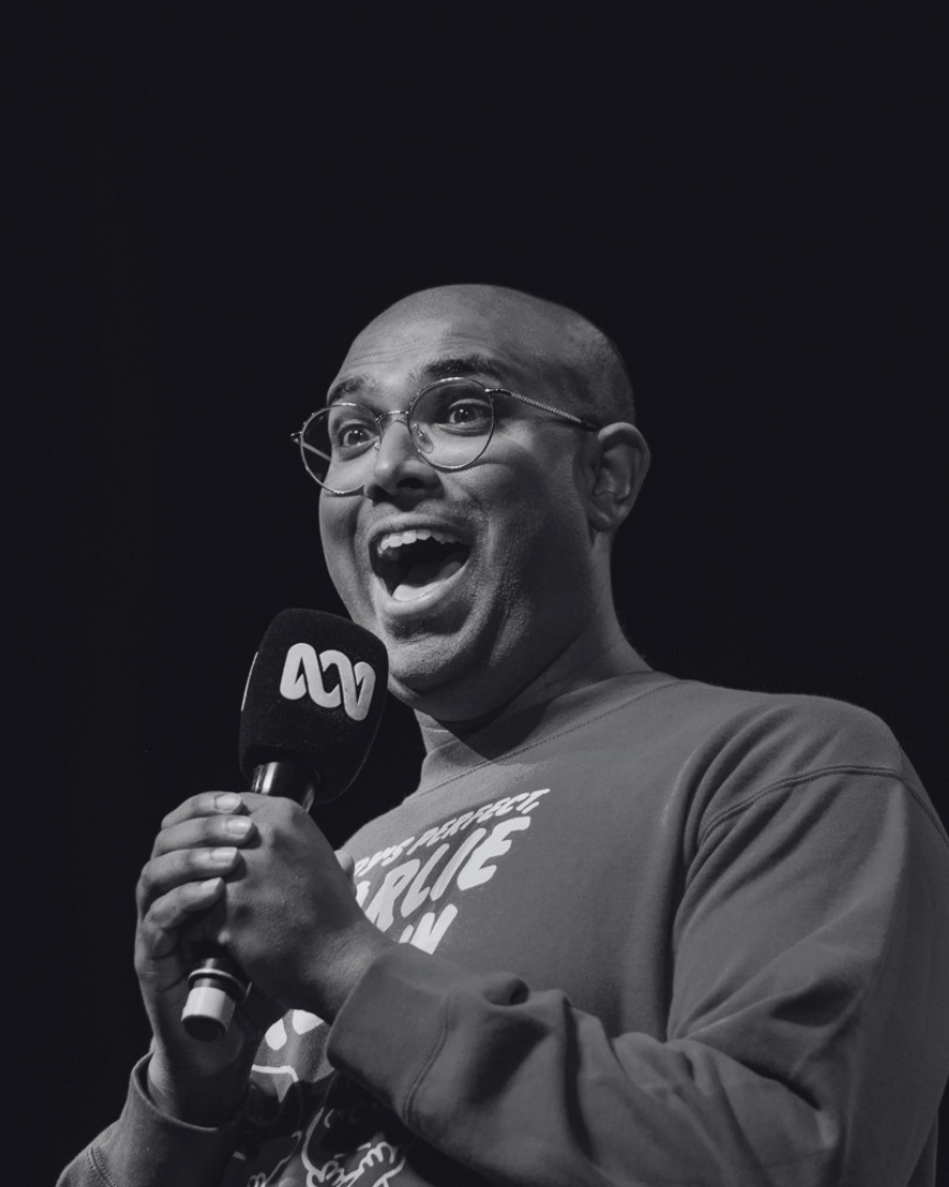 Rowan Thambar holding a microphone on a stage, smiling and speaking.