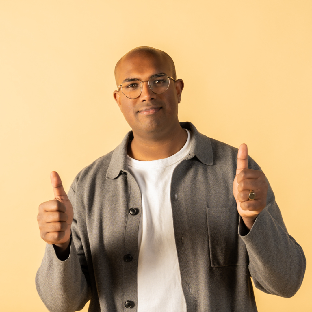 Rowan Thambar wearing glasses, a white t-shirt, and a gray jacket giving two thumbs up against a yellow background.