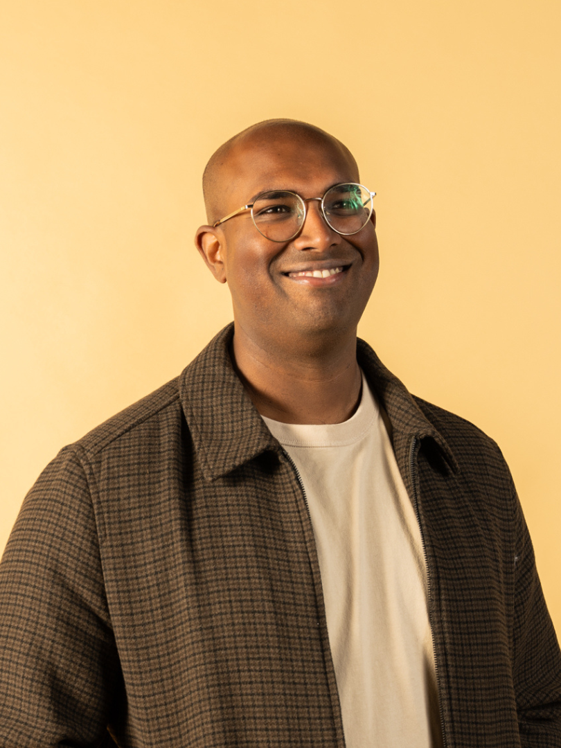 Rowan Thambar with glasses, wearing a collared jacket over a light-colored t-shirt, standing against a yellow background.