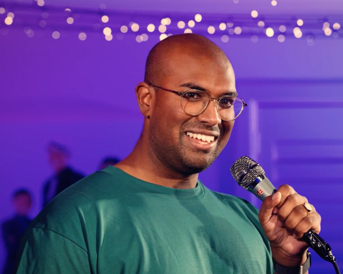 Rowan Thambar with glasses holding a microphone, smiling, in a setting with purple lighting and blurred background of people.