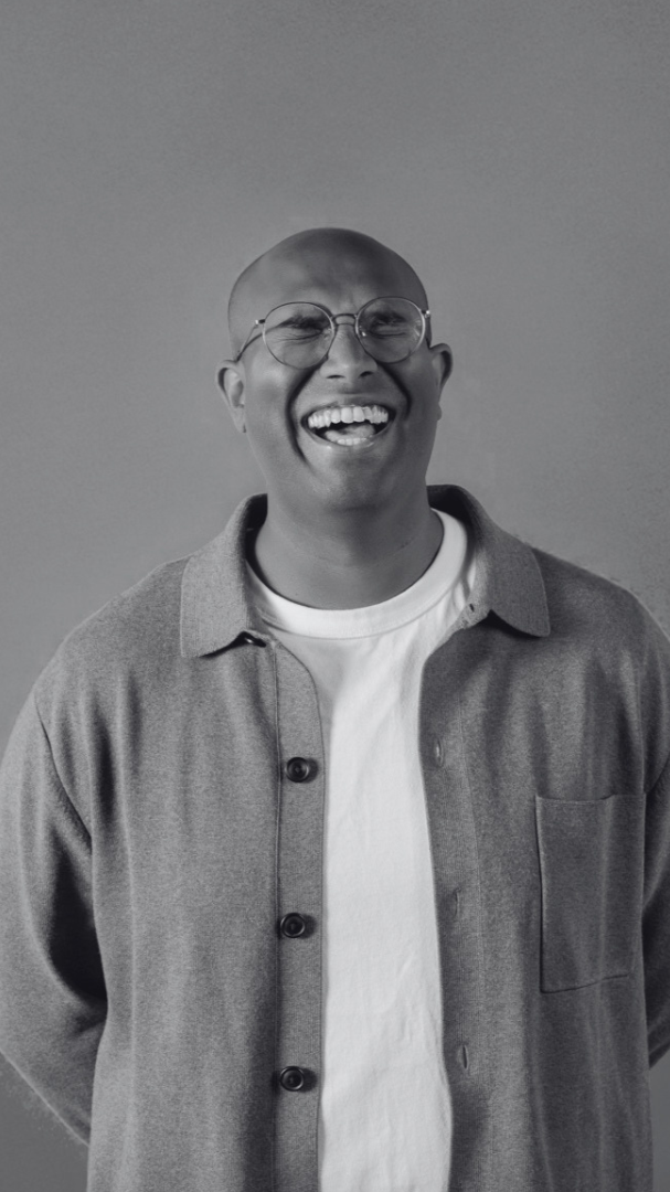 Rowan Thambar with round glasses, wearing a light-colored shirt and a darker jacket, standing against a plain background.