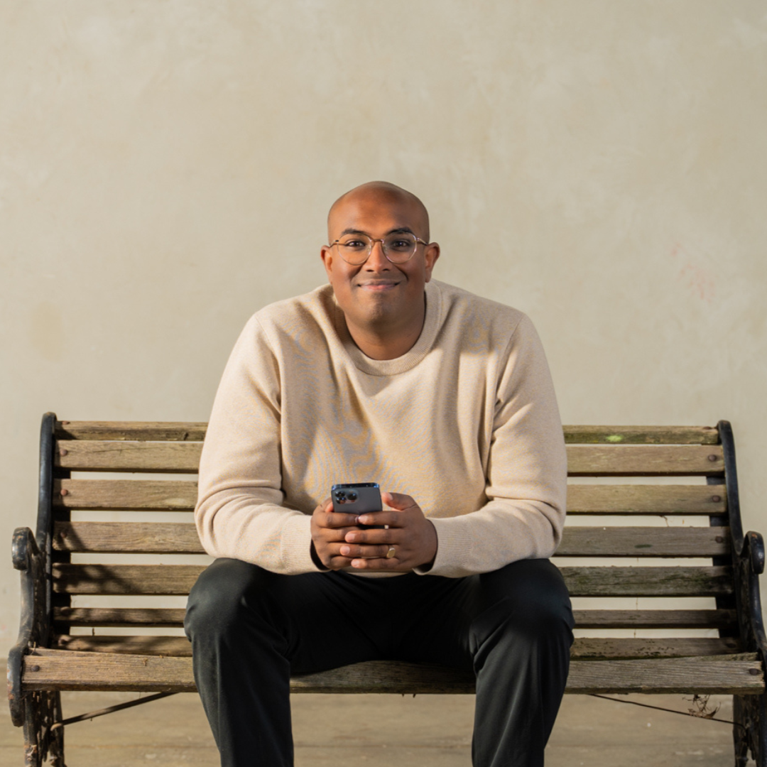 Rowan Thambar with glasses in a beige sweater sitting on a wooden bench holding a smartphone.