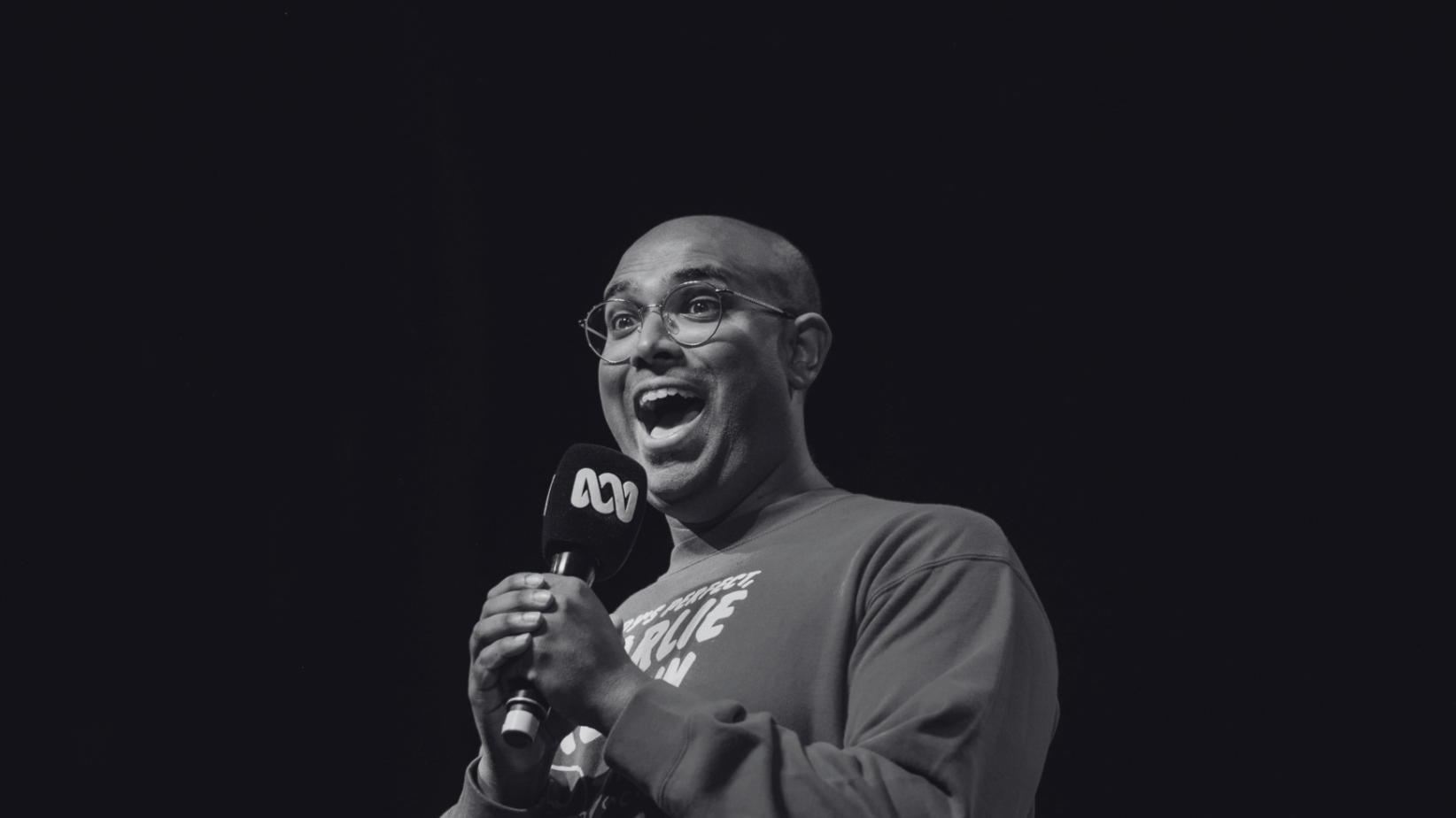 Rowan Thambar holding a microphone, smiling and speaking on stage with a dark background.
