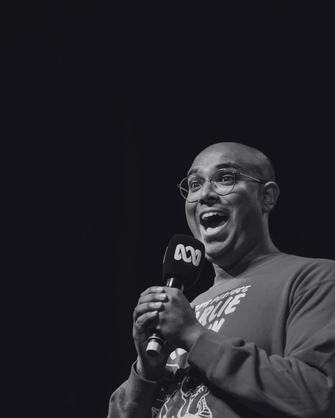 Rowan Thambar holding a microphone with an ABC logo, smiling and speaking, on stage in black-and-white.