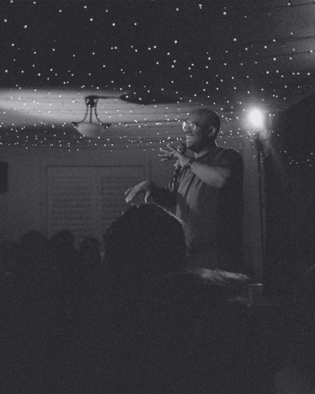 Rowan Thambar speaking or performing on stage with a microphone, underneath a ceiling decorated with small lights, in front of an audience in a dark room.