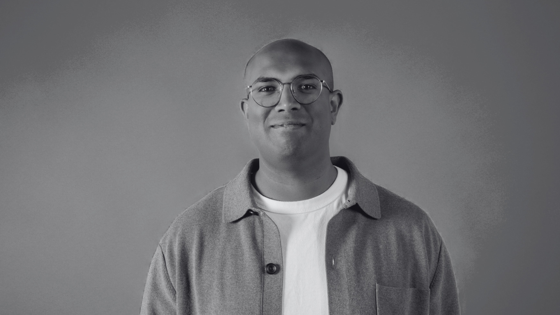 Black and white portrait of Rowan Thambar, short hair, wearing a white shirt and a gray jacket, smiling slightly against a plain background.