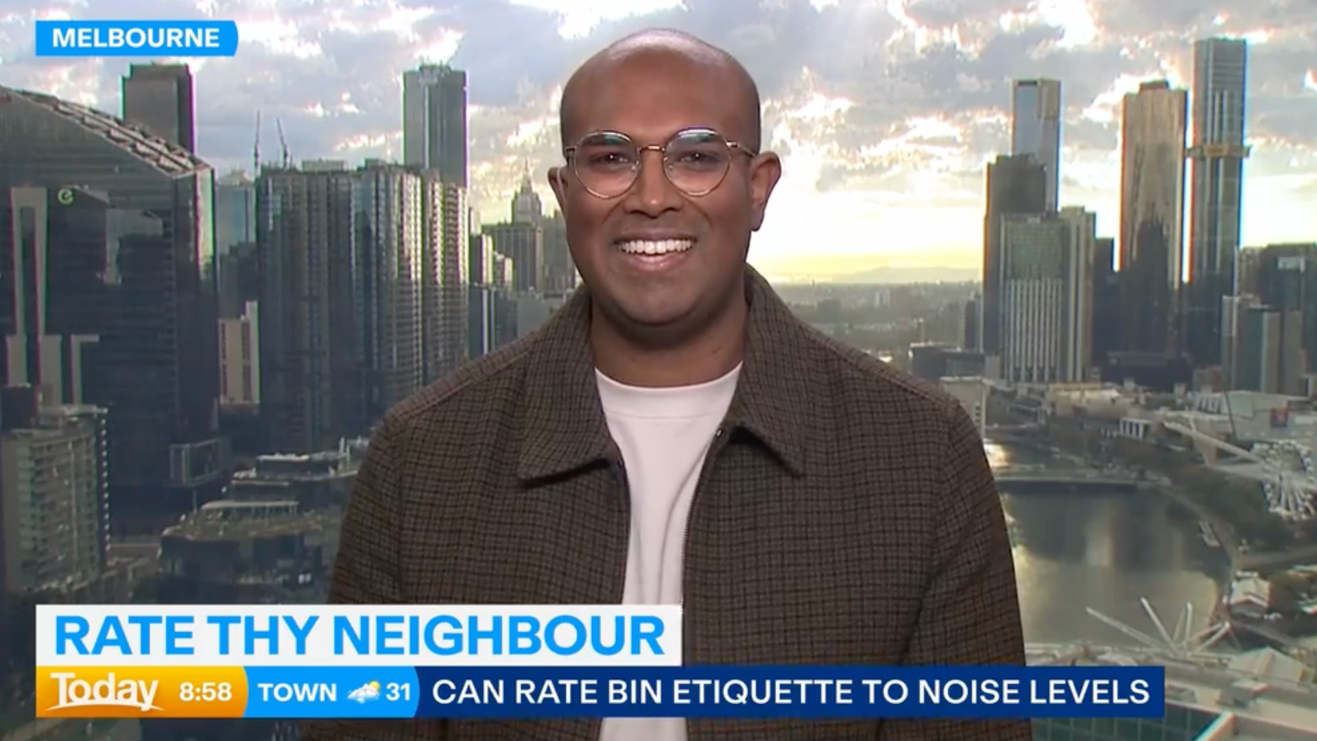 Rowan Thambar in brown checkered jacket smiling in front of a cityscape with tall buildings in Melbourne during daylight, with a river visible below. The video overlay includes the title 'Rate Thy Neighbour' and a news ticker mentioning noise levels.