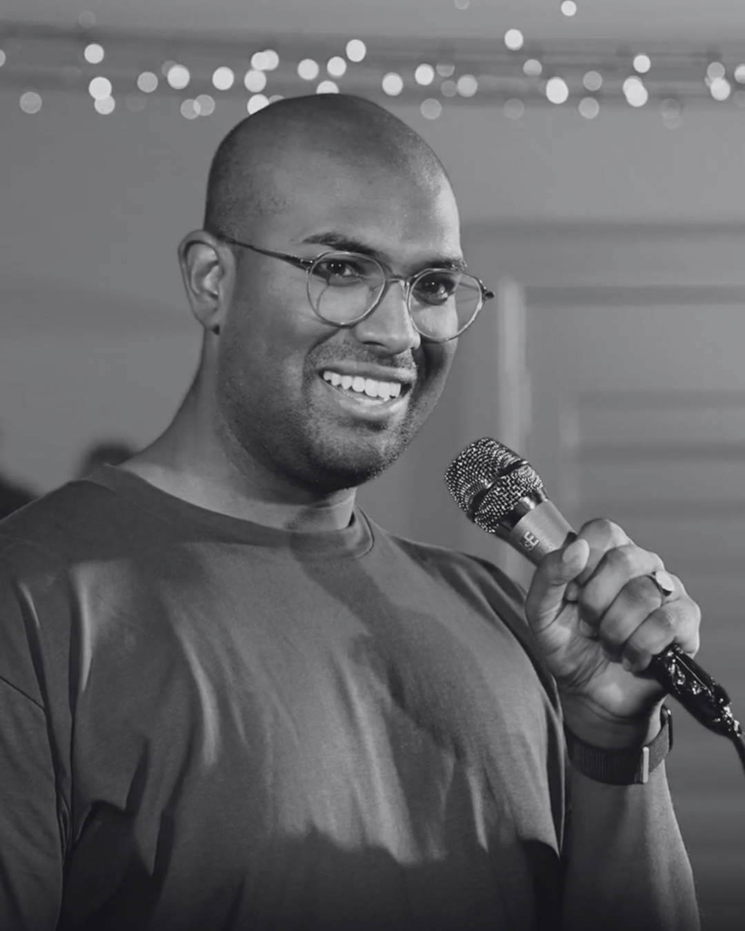Rowan Thambar holding a microphone indoors, with string lights in the background.