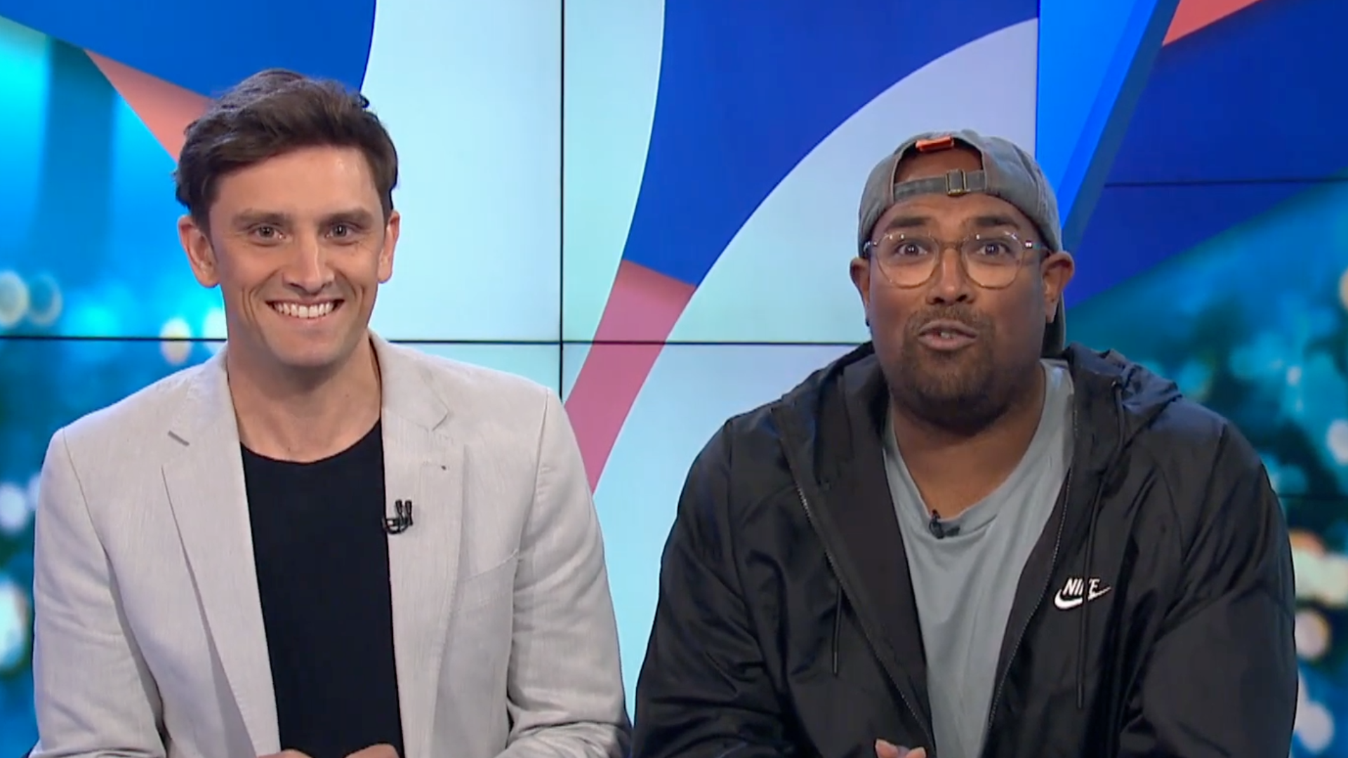 Two men sitting in front of a colorful television screen, with the man on the left wearing a light-colored blazer and a black shirt, and the man on the right wearing glasses, a backward cap, and a black Nike jacket.