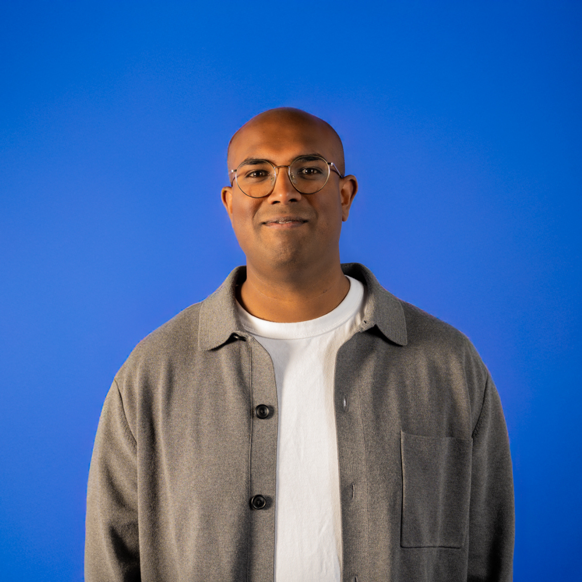 Portrait of a man with glasses, wearing a grey jacket over a white t-shirt, against a blue background.