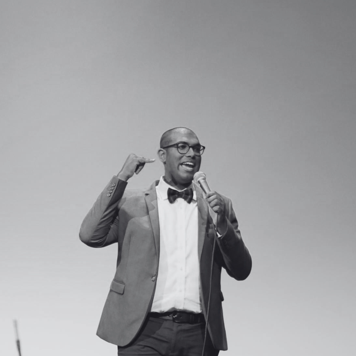 Rowan Thambar in a bow tie, dressed in a suit, holding a microphone, speaking or performing on stage.