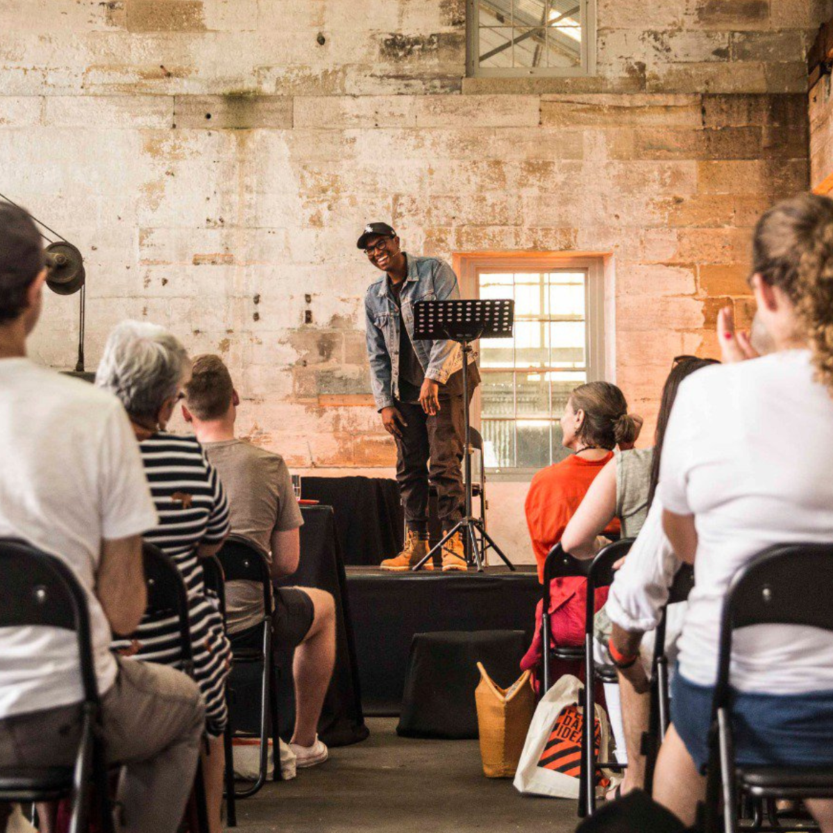 Rowan Thambar standing on a stage giving a presentation to an audience in a rustic room with exposed brick walls and large windows.