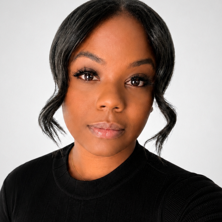 Close-up portrait of a woman with dark hair styled in loose waves, wearing a black top, with a neutral expression against a plain light gray background.