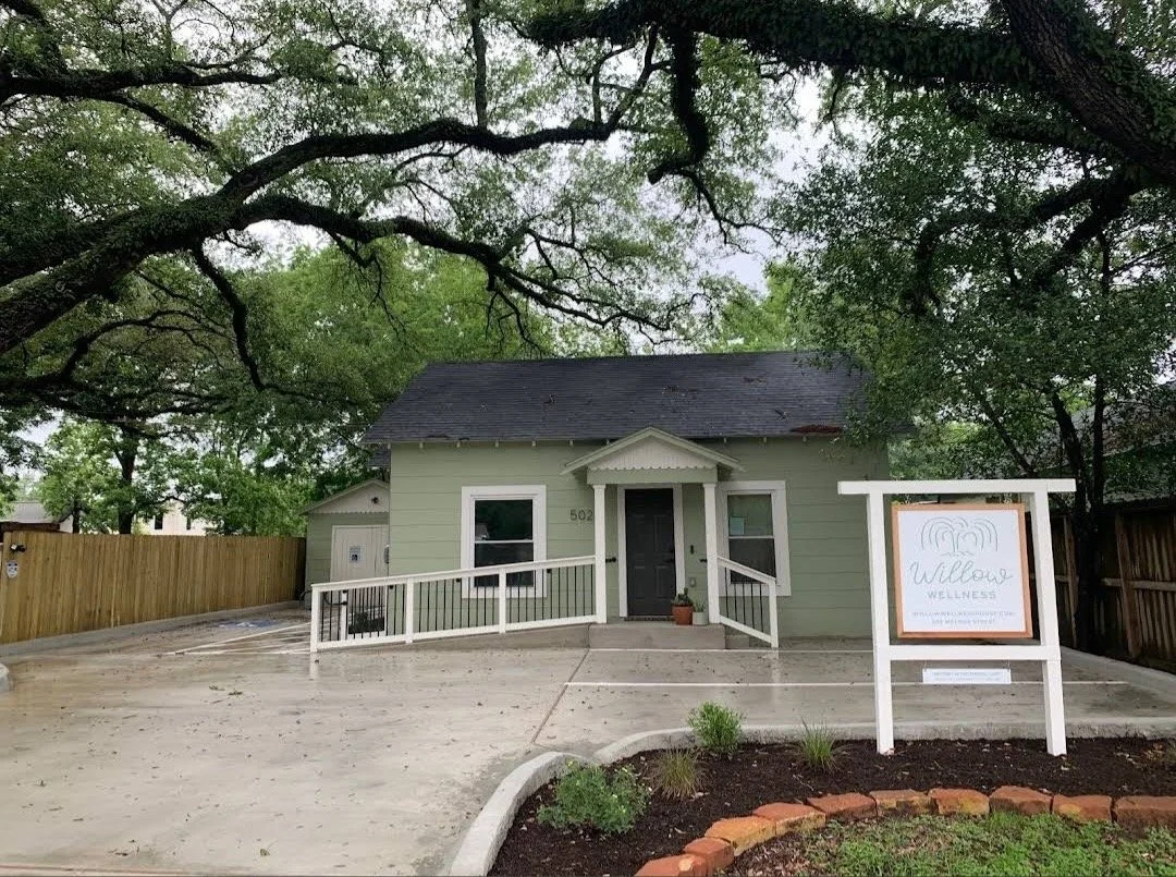 Affordable EMDR Therapy in the heart of Old Tomball, TX. Exterior of a small, light green wellness clinic with a black door, sign that reads 'Willow Wellness,' and a concrete walkway leading to the entrance, surrounded by trees.