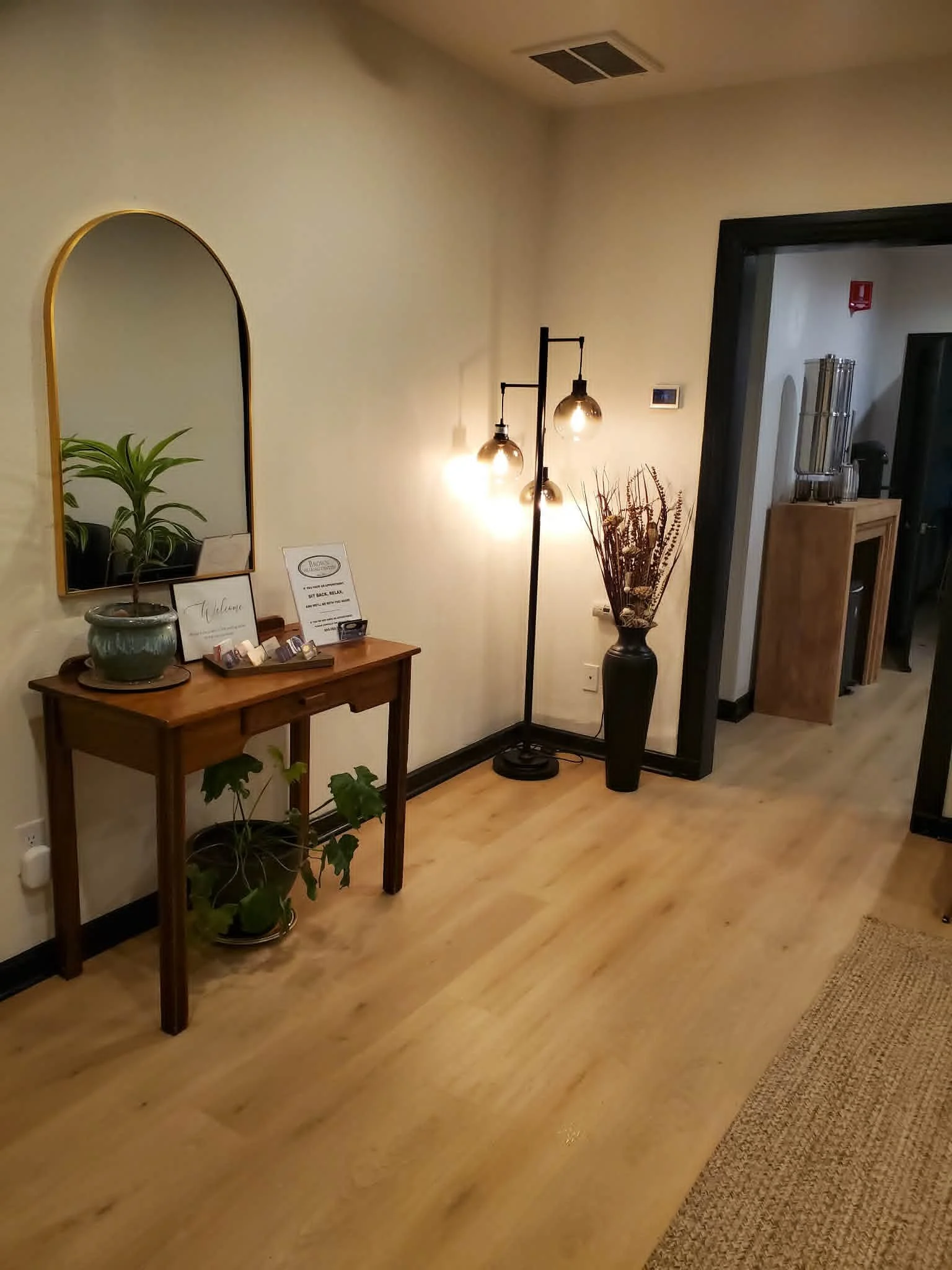 An interior room corner with a small wooden table holding a potted plant, framed signs, and small items. Next to the table is a tall black floor lamp with three globe lights, and a tall vase with decorative dried branches. On the wall, a mirror reflects the plant and table. Parts of a doorway and a beverage station are visible in the background.