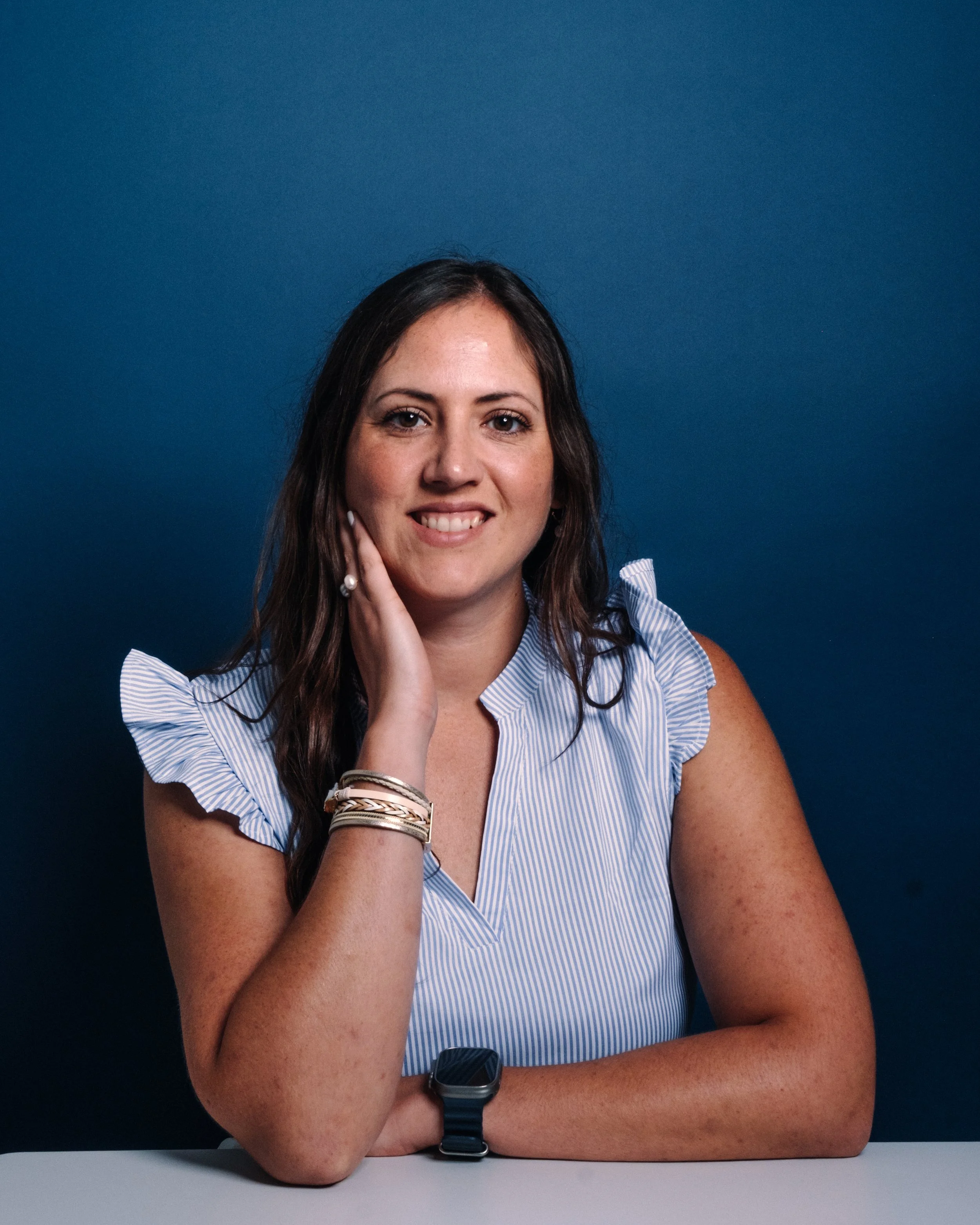 A young woman with long brown hair, wearing a blue and white striped sleeveless blouse, leather bracelets, and a smartwatch, smiling and resting her chin on her hand against a solid blue background.