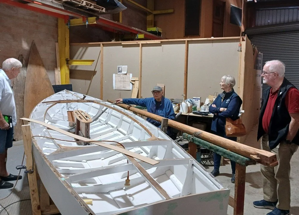 Members and guests observe 'Falcon III' under renovation. The boat is on a workbench, with building materials and tools around.