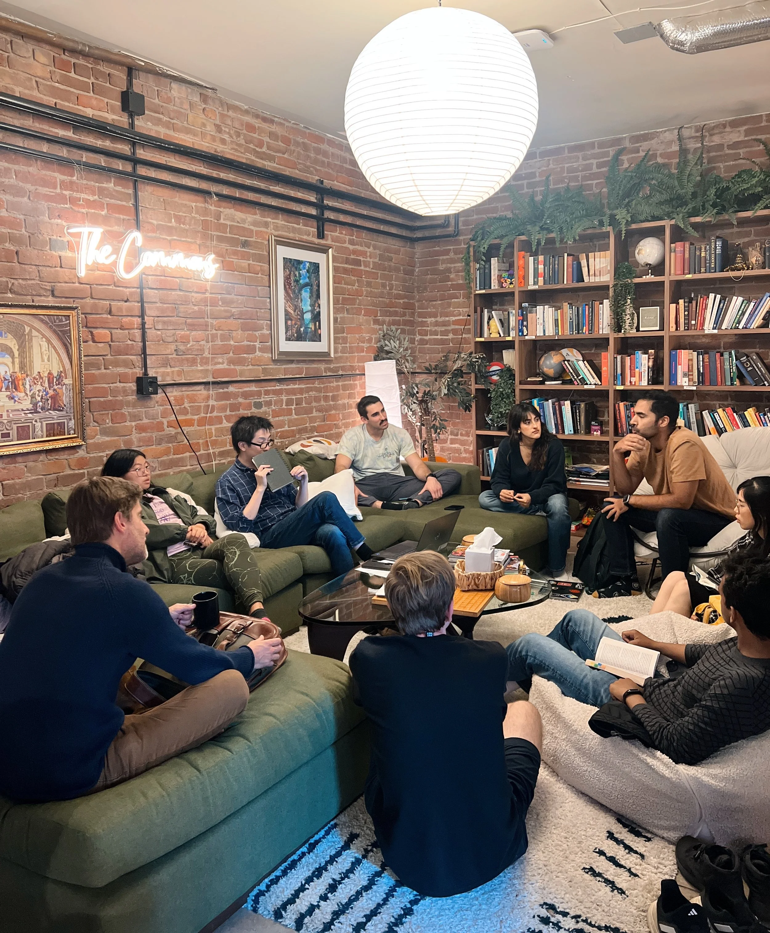A group of young adults sitting in a cozy living room with exposed brick walls, a large paper lantern ceiling light, a bookcase filled with books and decorative items, and a patterned rug. Some people are sitting on a green sofa and others on the floor, engaged in conversation.