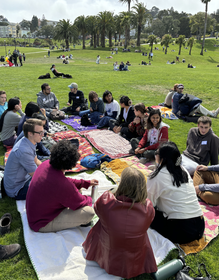 Group of people sitting on blankets in a park, engaging in conversation, with palm trees and a grassy area in the background.