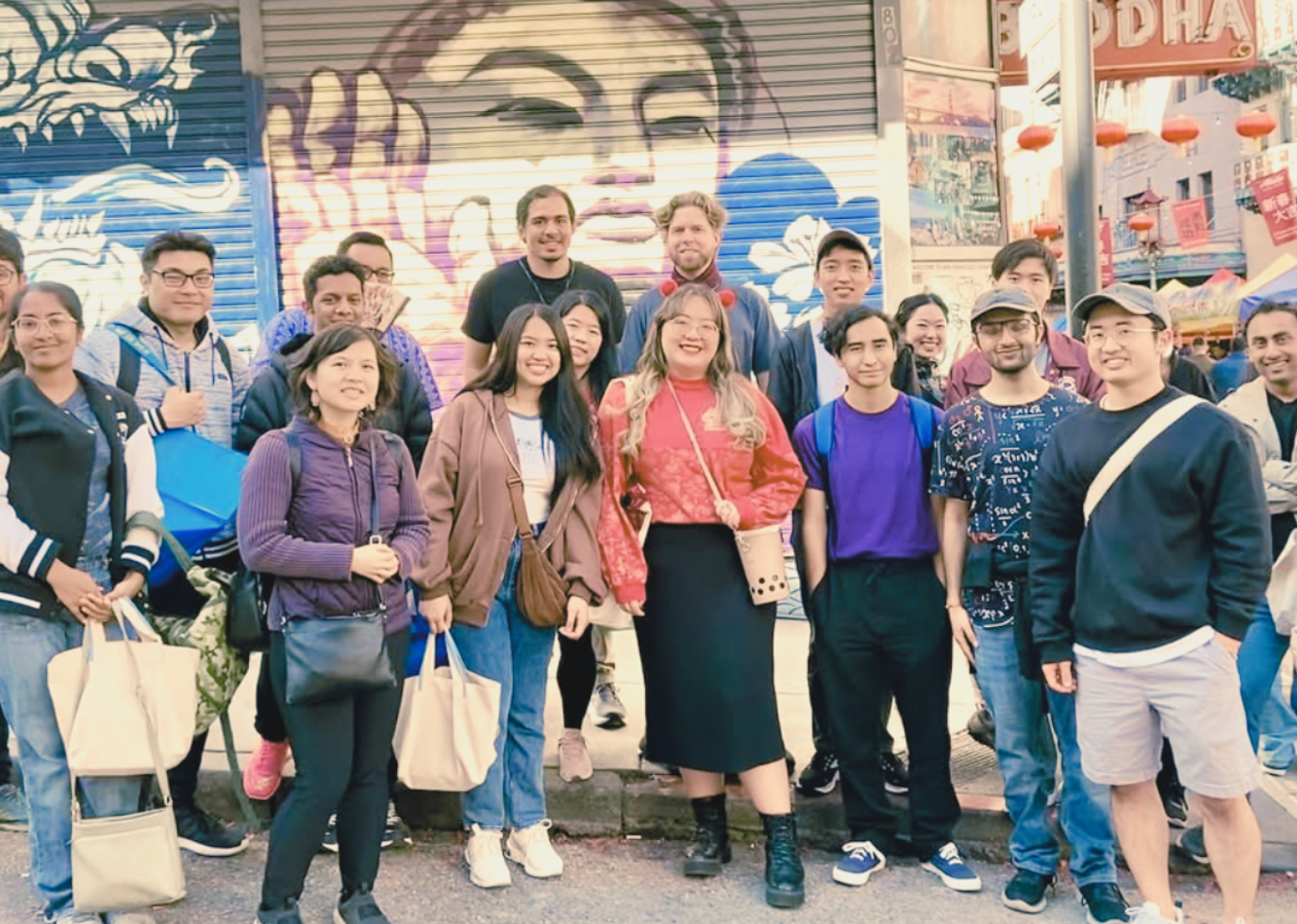 A group of diverse young adults smiling and posing for a photo on a city street with colorful graffiti art in the background.