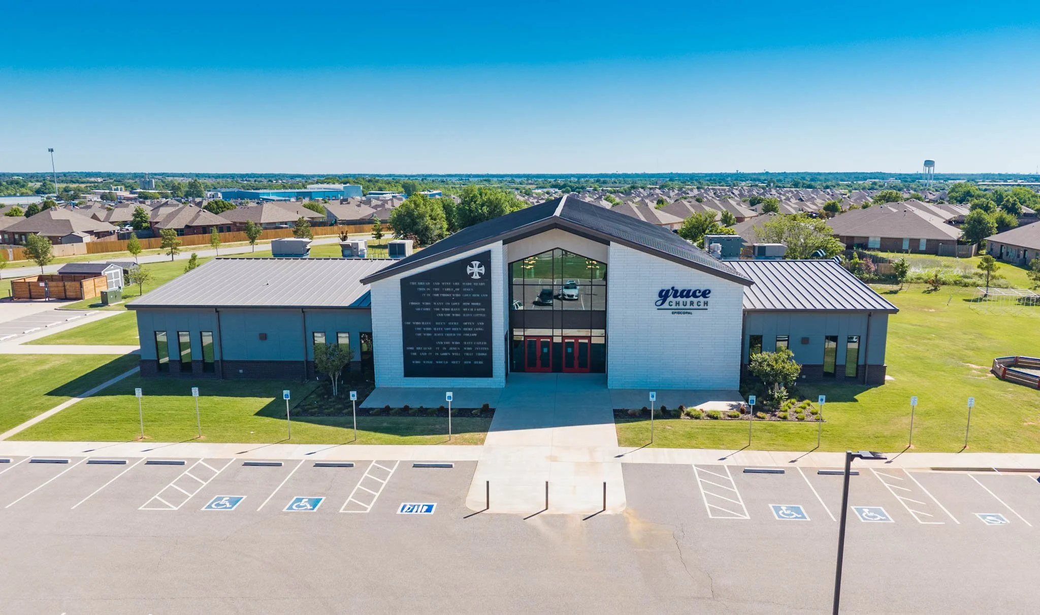 Aerial view of Grace Church Episcopal with a parking lot marked for handicapped spaces in the foreground, green grass, trees, residential houses, and a clear blue sky in the background.