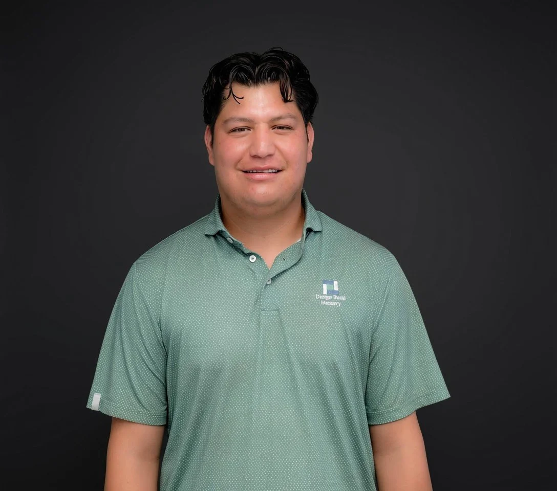 A man with dark hair smiling in front of a dark grey background, wearing a green polo shirt with a logo.