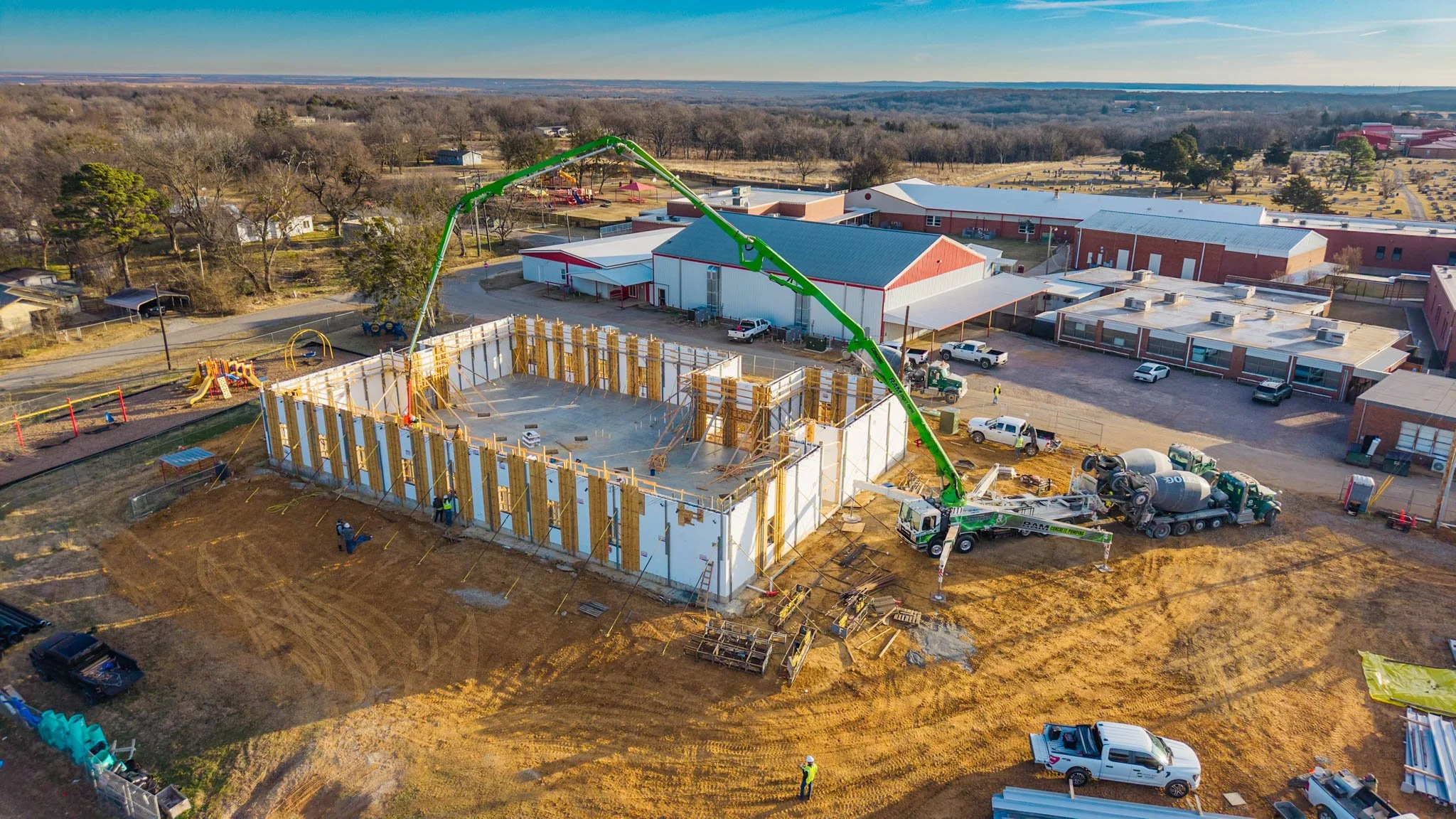 An aerial view of a construction site with a partially built structure, a large green crane, several construction workers, and surrounding vehicles. In the background, there are buildings, trees, and a rural landscape.