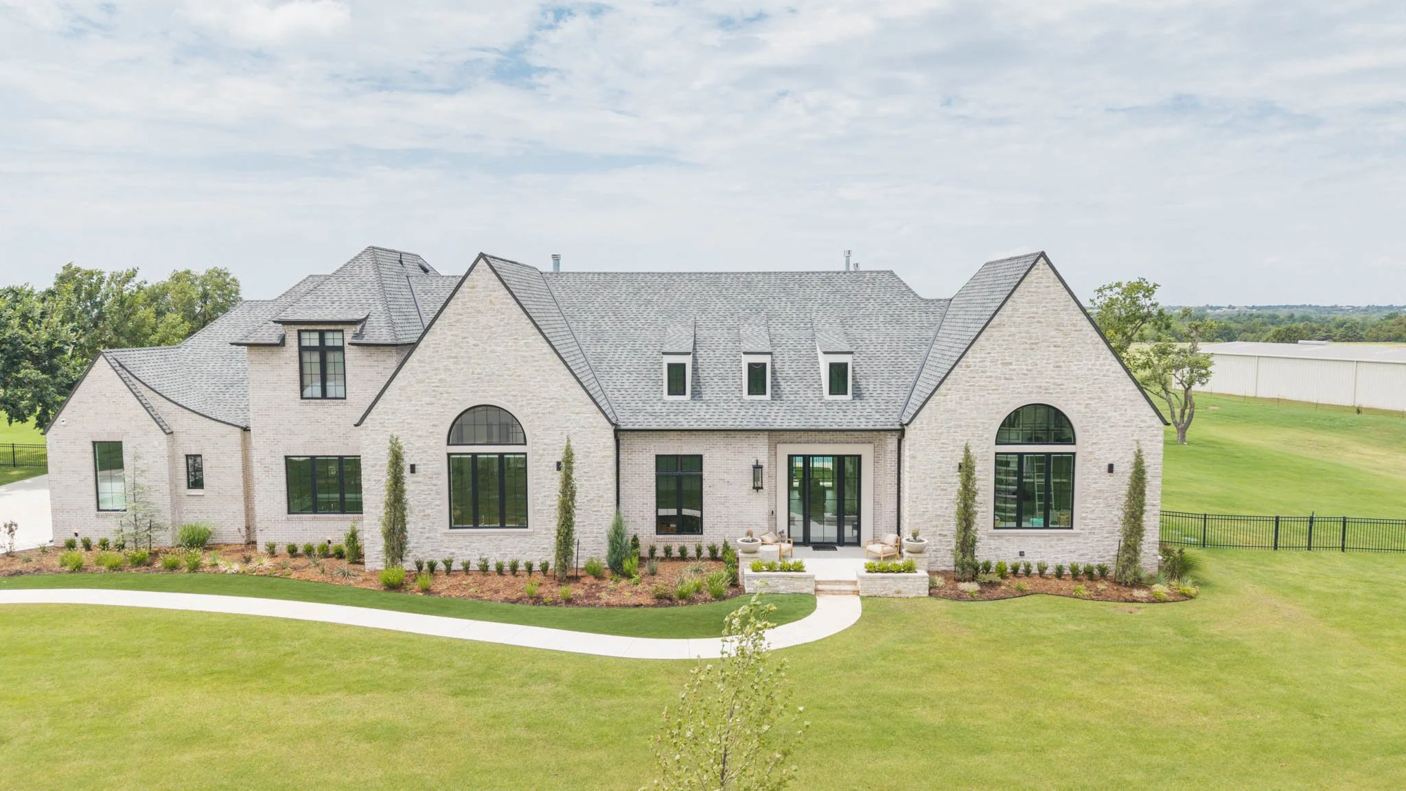 Front view of a large, modern house with a gray slate roof, white brick exterior, and large arched windows, surrounded by a well-maintained green lawn and landscaped garden.