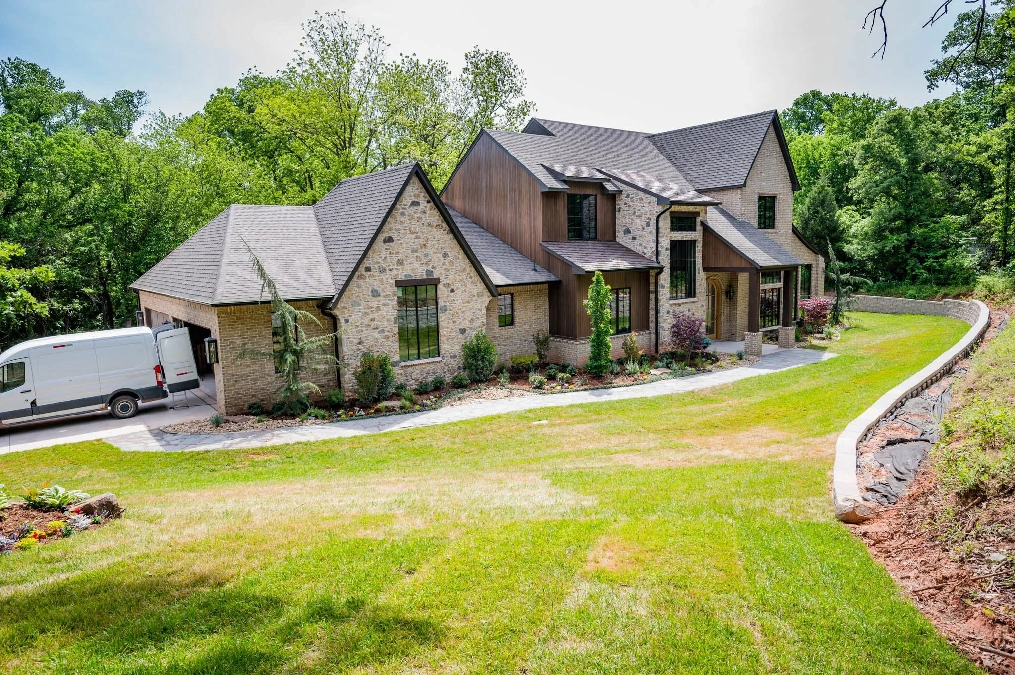 A large modern house with stone and wood exterior, surrounded by green trees and a well-maintained lawn, with a white van parked in the driveway.
