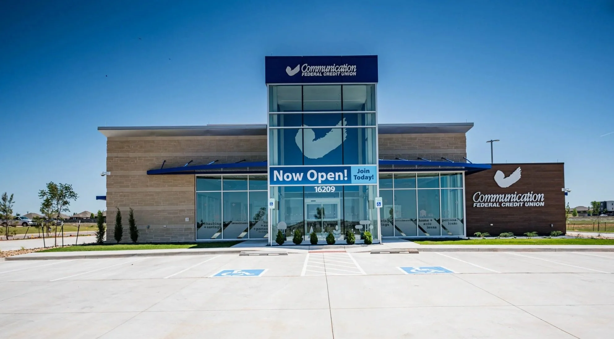Newly opened Communication Federal Credit Union branch with a sign saying 'Now Open! Join Today!' located at 16209, with parking spaces, including designated handicap spots, and a clear blue sky.