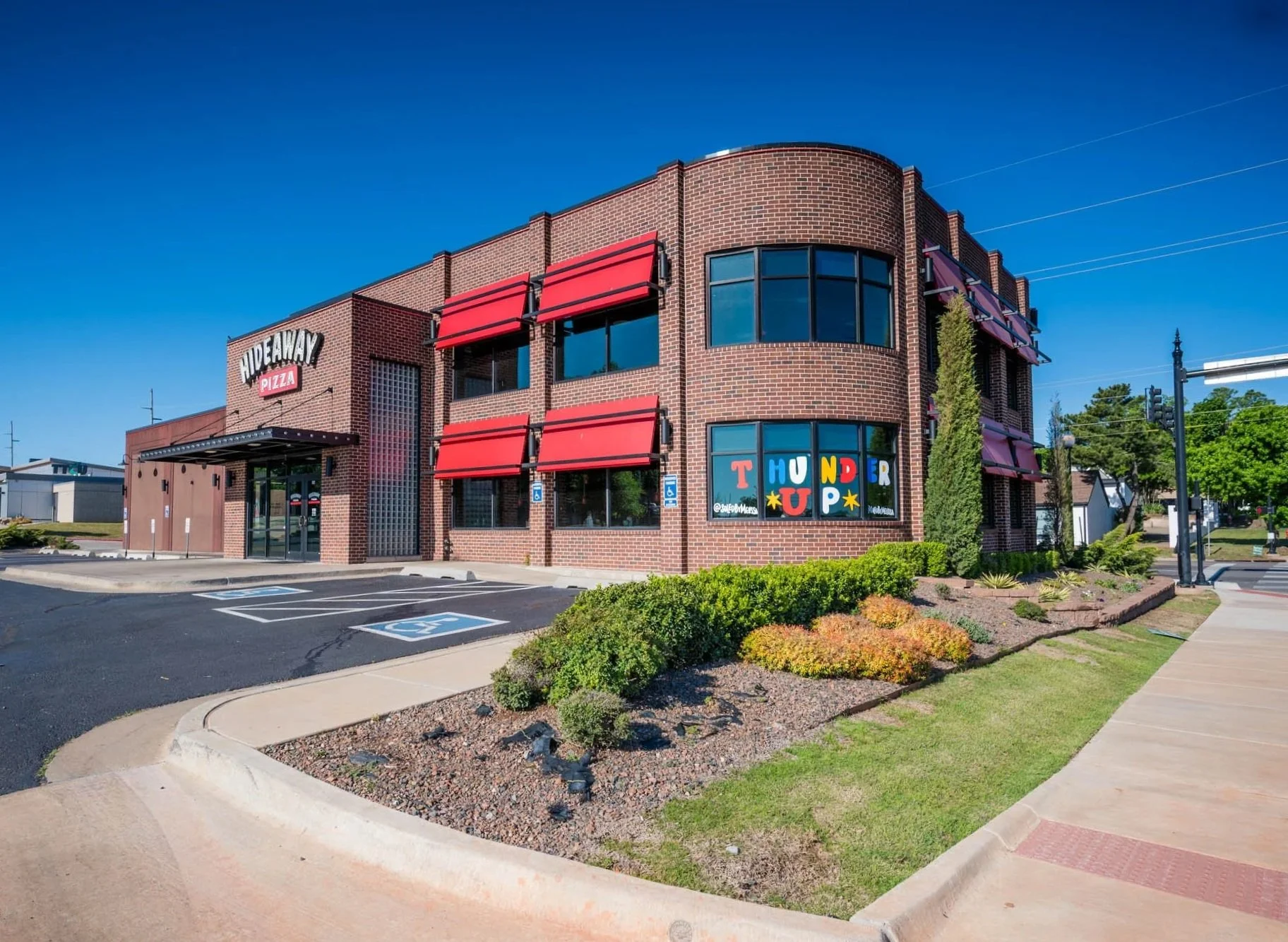 A two-story brick building with red awnings and large windows, with a sign that reads "Hideaway Pizza." The parking lot in front has designated handicapped spaces, and there is a garden bed with bushes and plants along the sidewalk.
