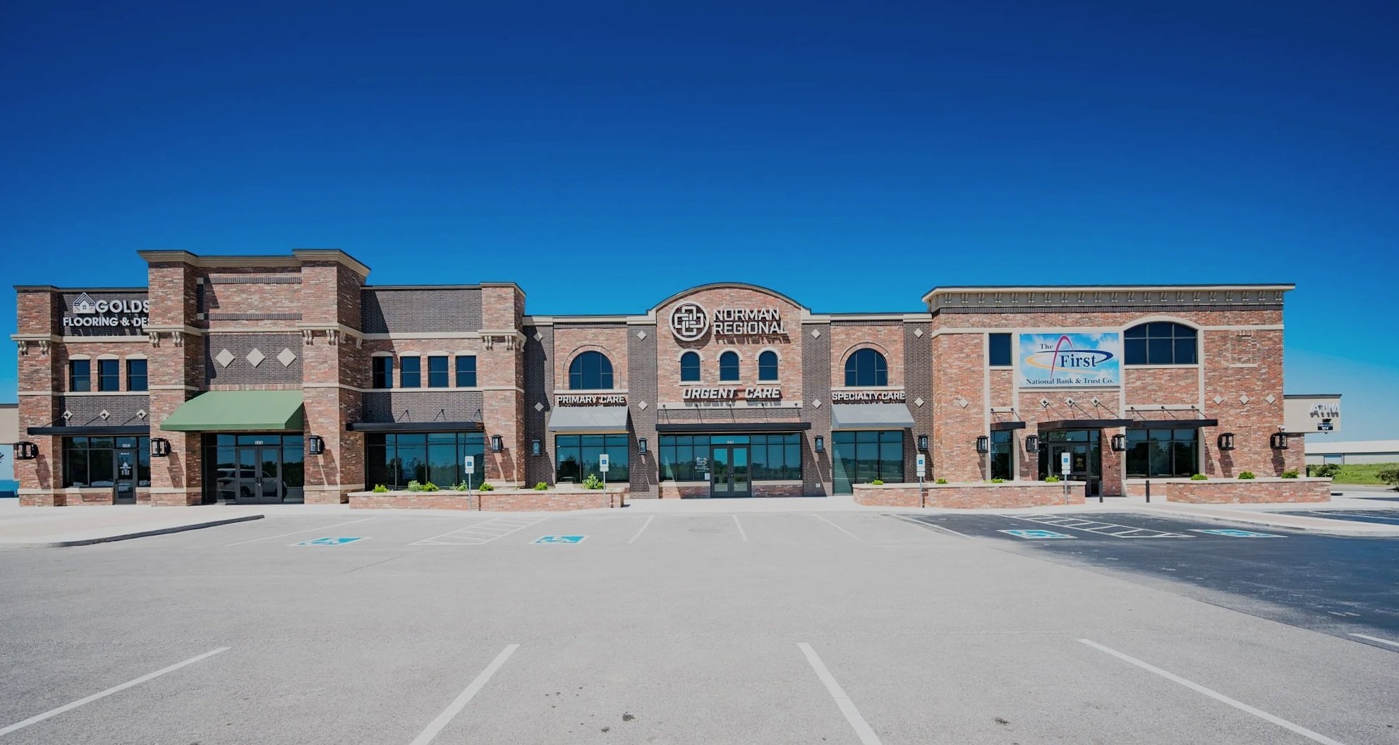 A brick medical office building with signs for Norman Regional, primary care, urgent care, and specialty care. The parking lot is empty with marked spaces, including handicapped spots near the entrance. The sky is clear and blue.
