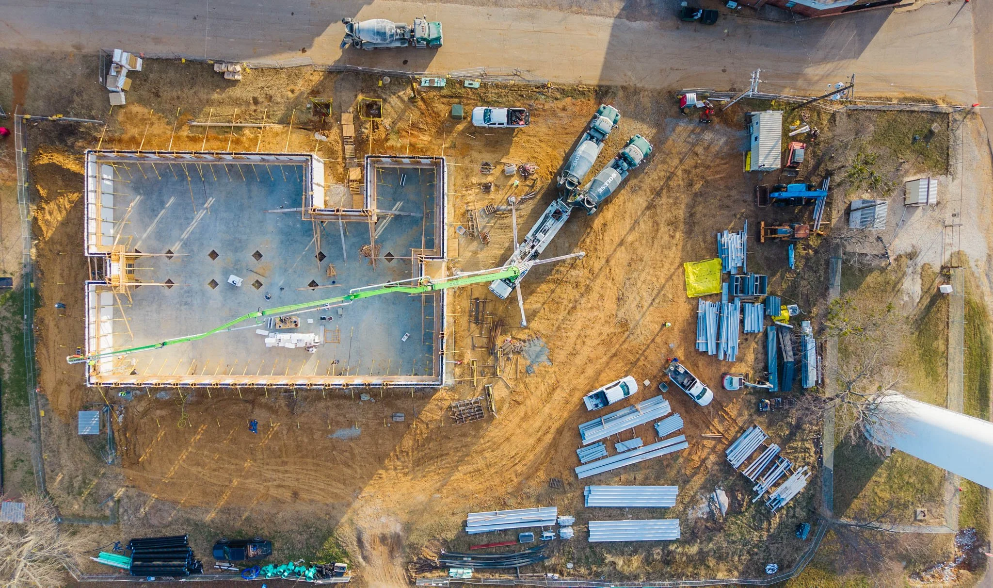 An aerial view of a construction site showing building foundations, construction equipment, trucks, scaffolding, and materials, with areas of dirt and partially built structures.