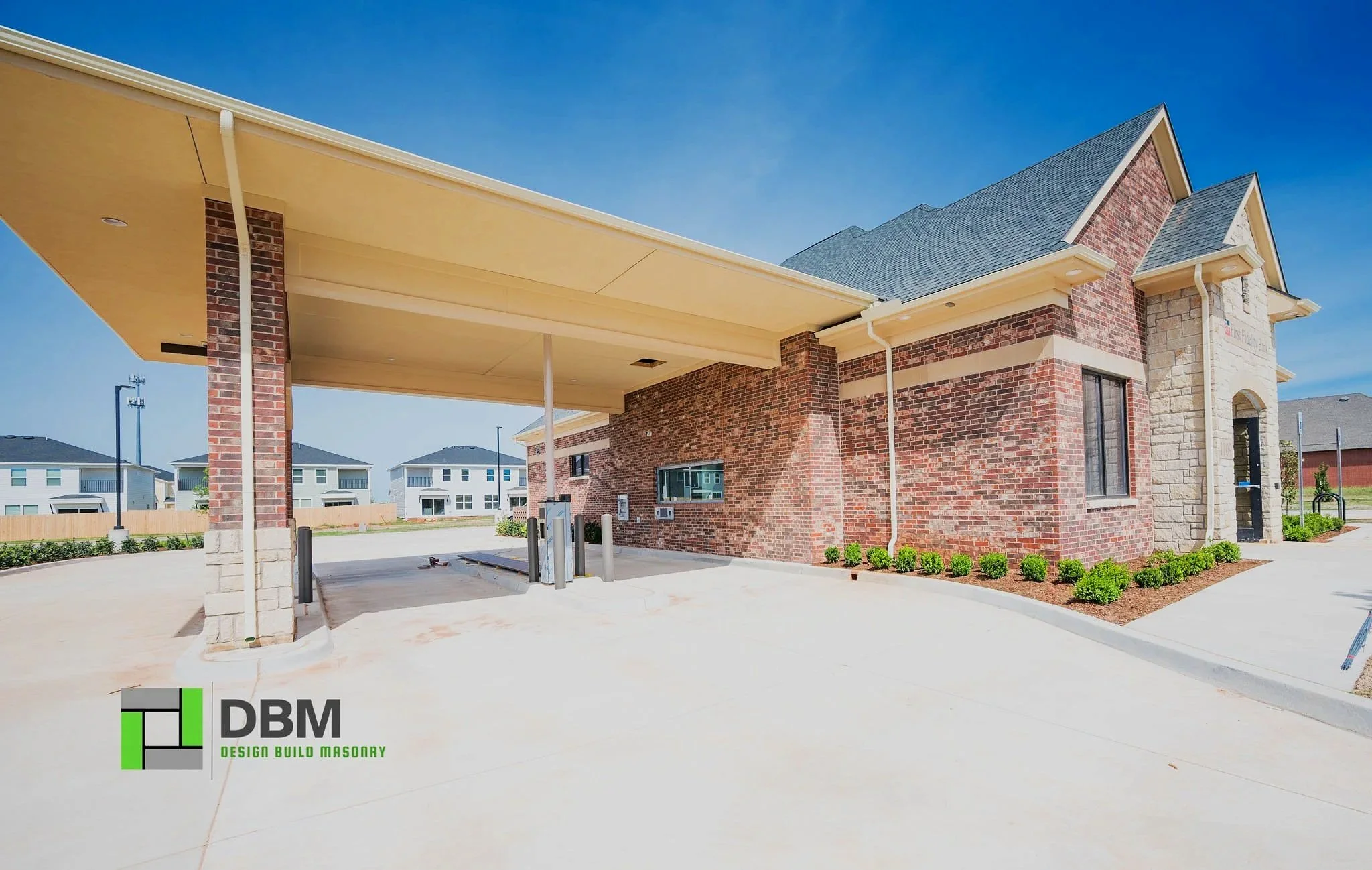 Newly constructed brick building with a covered parking area and landscaped shrubs in front, under a clear blue sky.