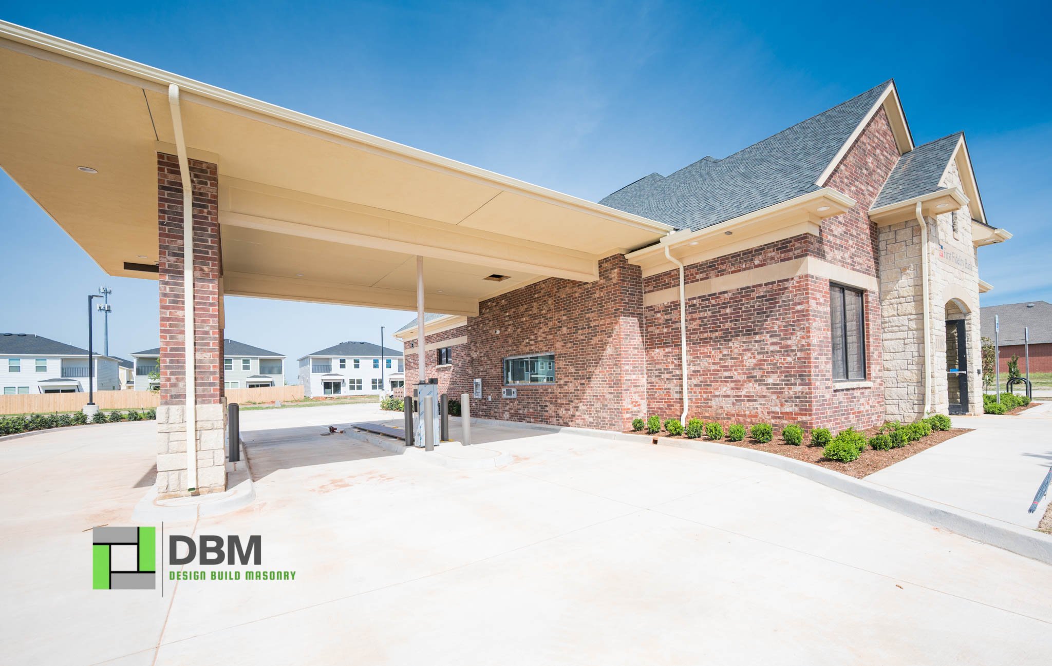 A new brick building with a covered parking area supported by brick pillars, surrounded by small green bushes and a concrete parking lot, under a clear blue sky.