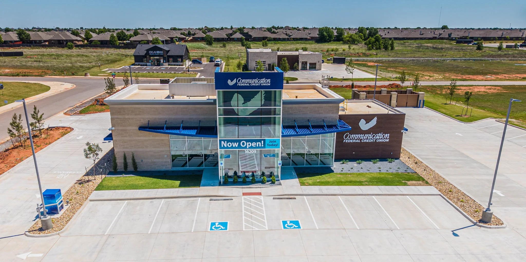 Aerial view of a new commercial building for Communication Federal Credit Union with a sign indicating it is now open. The building has large glass windows and parking spaces, including designated accessible spots, with a mostly empty parking lot. Surrounding the building are small trees and lampposts, with a residential neighborhood visible in the background.