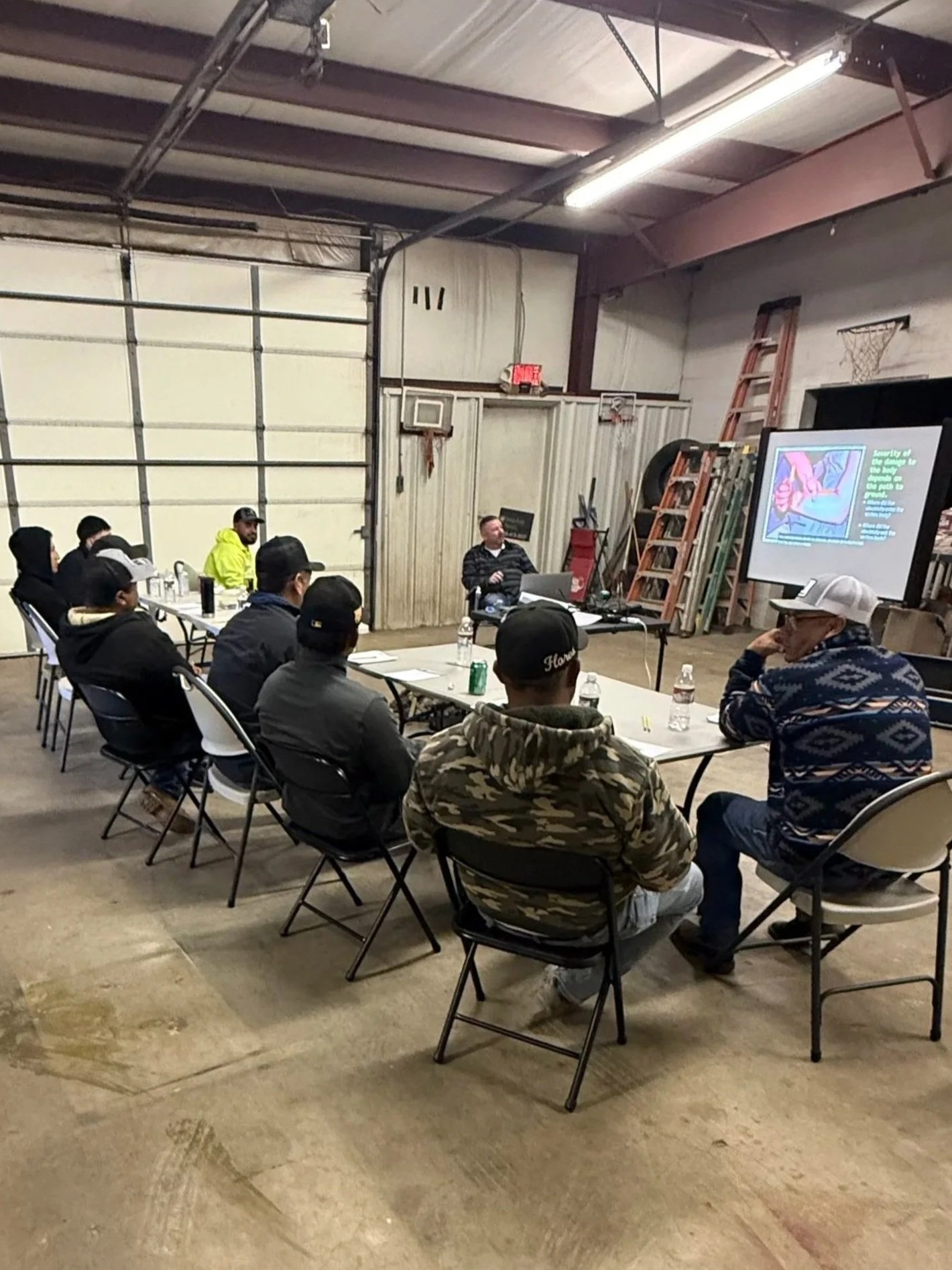 Group of people seated around a table in an industrial or warehouse setting listening to a presenter. The presenter is on the right, seated at a table with a laptop, and a projector screen displays a presentation. The room has a high ceiling, exposed beams, and equipment like ladders and storage areas.