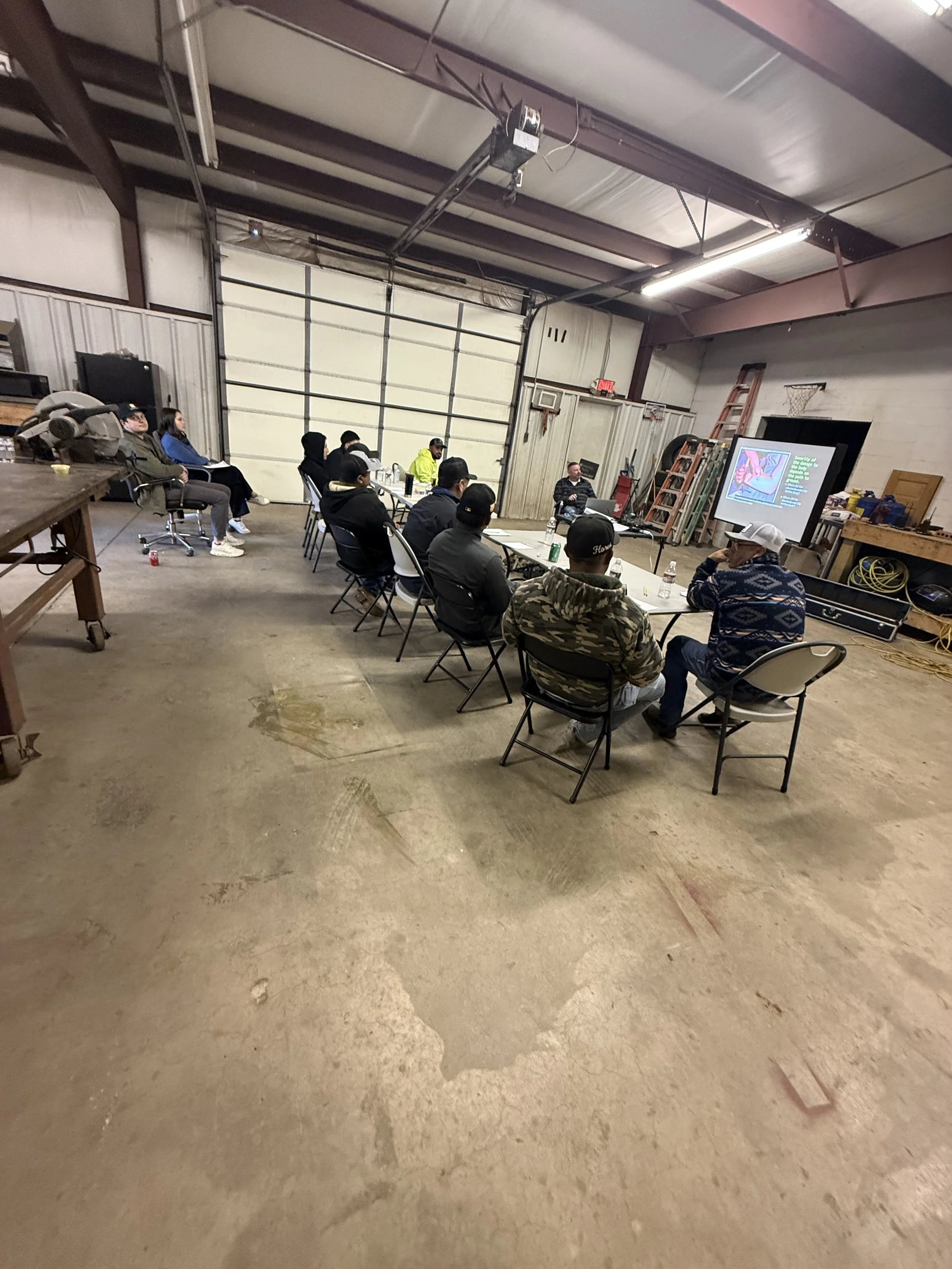 Group of people sitting at a long table in a warehouse or garage setting, attending a presentation with a person speaking near a large screen at the front.
