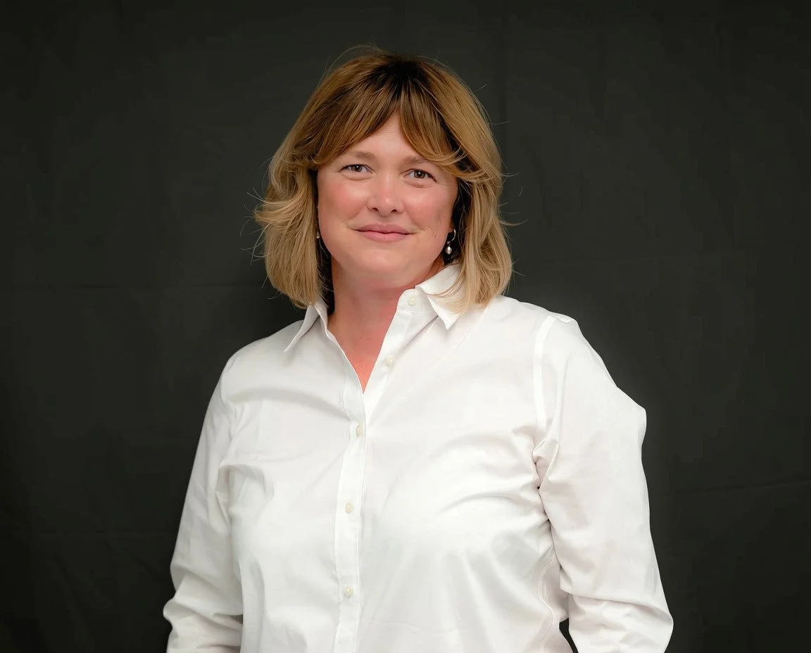 A woman with shoulder-length blonde hair wearing a white button-down shirt, standing against a dark background.