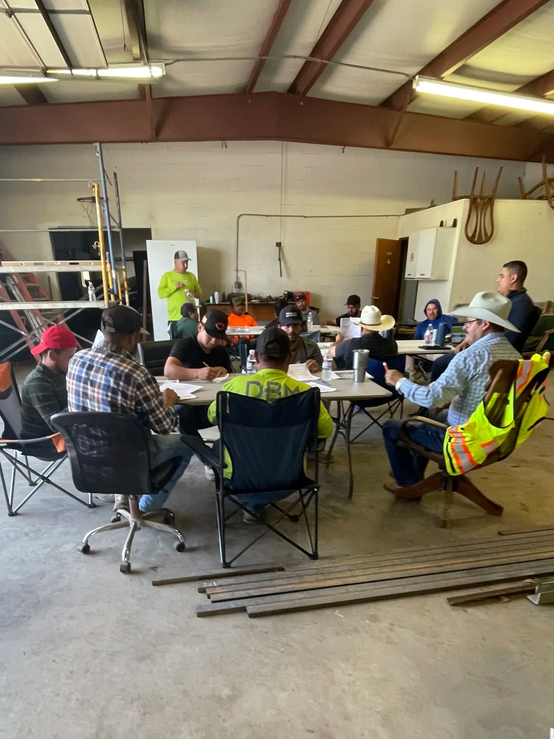 A group of people gathered around tables inside a warehouse or workshop, some wearing cowboy hats and safety vests. A man in a bright green shirt is standing and speaking to the group. Various equipment and materials are around, including a ladder, chairs, and a wooden pallet in the foreground.