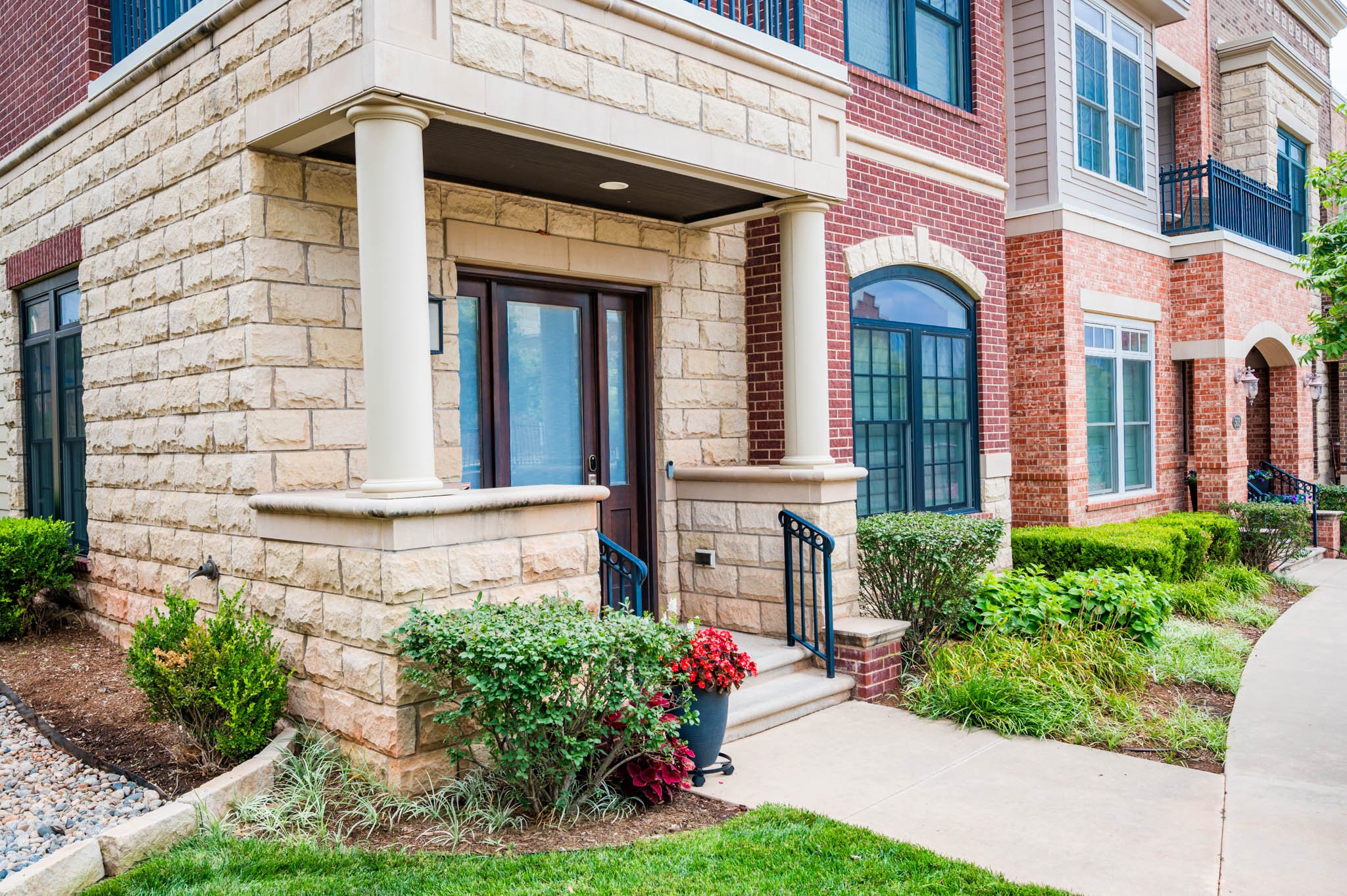 Front exterior of a modern apartment building with brick and stone facade, featuring large windows, a small porch with white columns, a concrete sidewalk, landscaped bushes, and a small garden area.