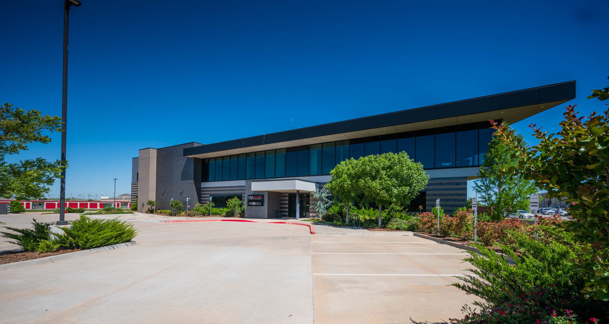Modern commercial building with large glass windows and landscaping, situated in a parking lot with trees and shrubbery, under a clear blue sky.