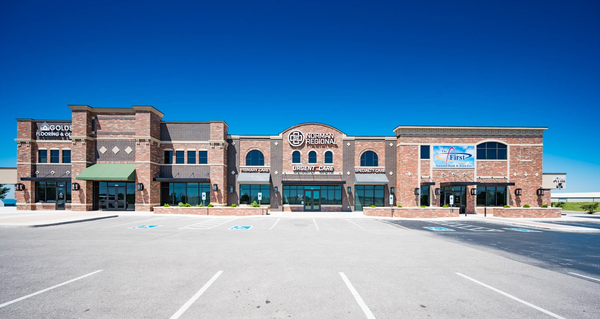 A brick medical office building with signs for Norman Regional, primary care, urgent care, and specialty care. The parking lot is empty with marked spaces, including handicapped spots near the entrance. The sky is clear and blue.