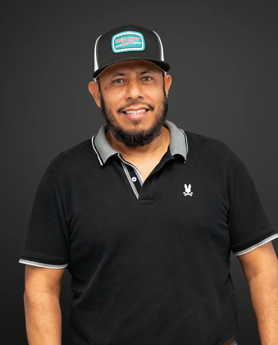 A man with a beard smiling, wearing a black polo shirt with white accents and a logo, and a black cap with a colorful patch, standing against a dark background.