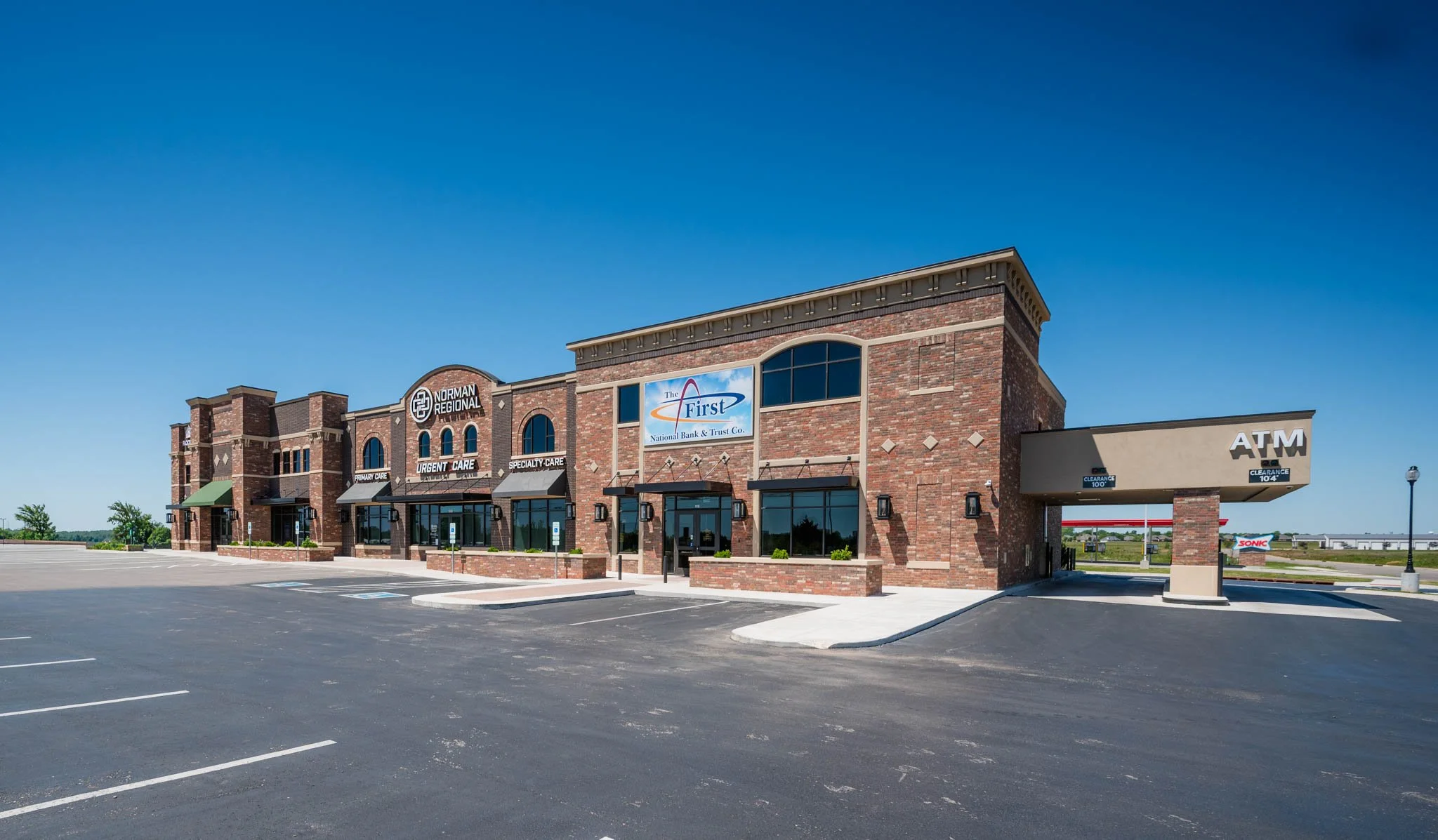 A strip mall with a brick facade, featuring stores such as Norman Regional, urgent care, specialty care, and The First bank. There is a parking lot in front with marked parking spaces, and a clear blue sky overhead.