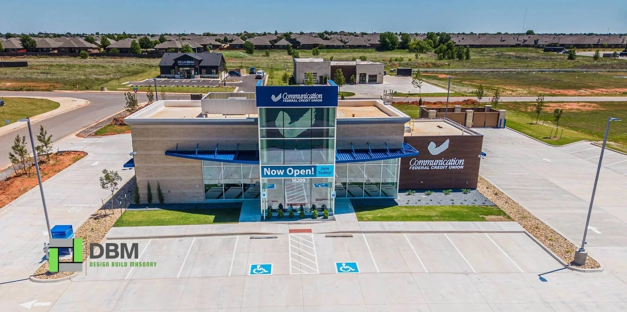 A new Communication Federal Credit Union branch with a sign that reads "Now Open! Join Today!" in a parking lot. The building has large glass windows and a modern design, with nearby parking spaces and trees, and is situated in a suburban area with other commercial buildings and houses in the background.