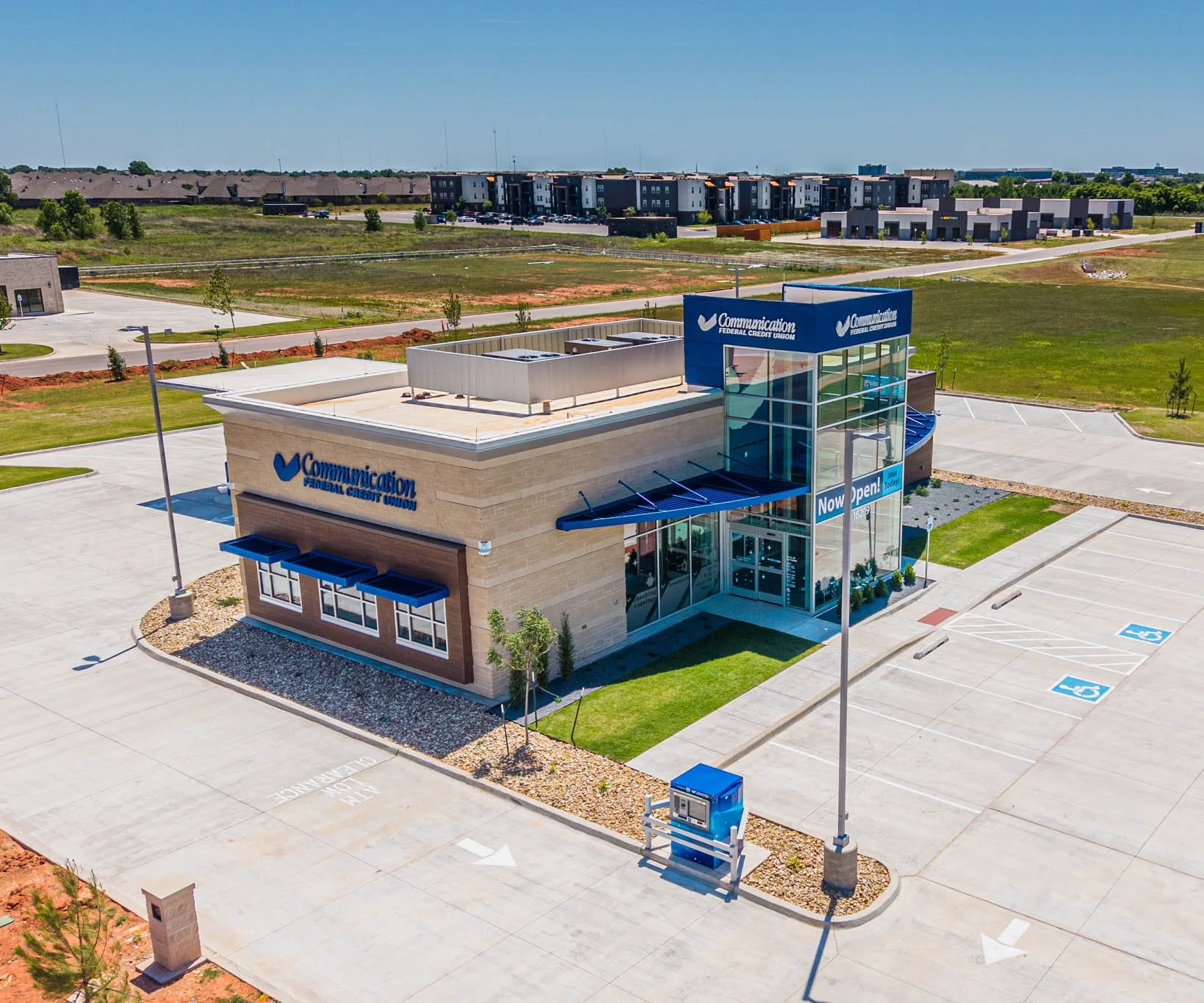 An empty parking lot outside a new Communication Federal Credit Union branch with a 'Now Open!' sign, surrounded by a grassy area and other residential buildings in the background.