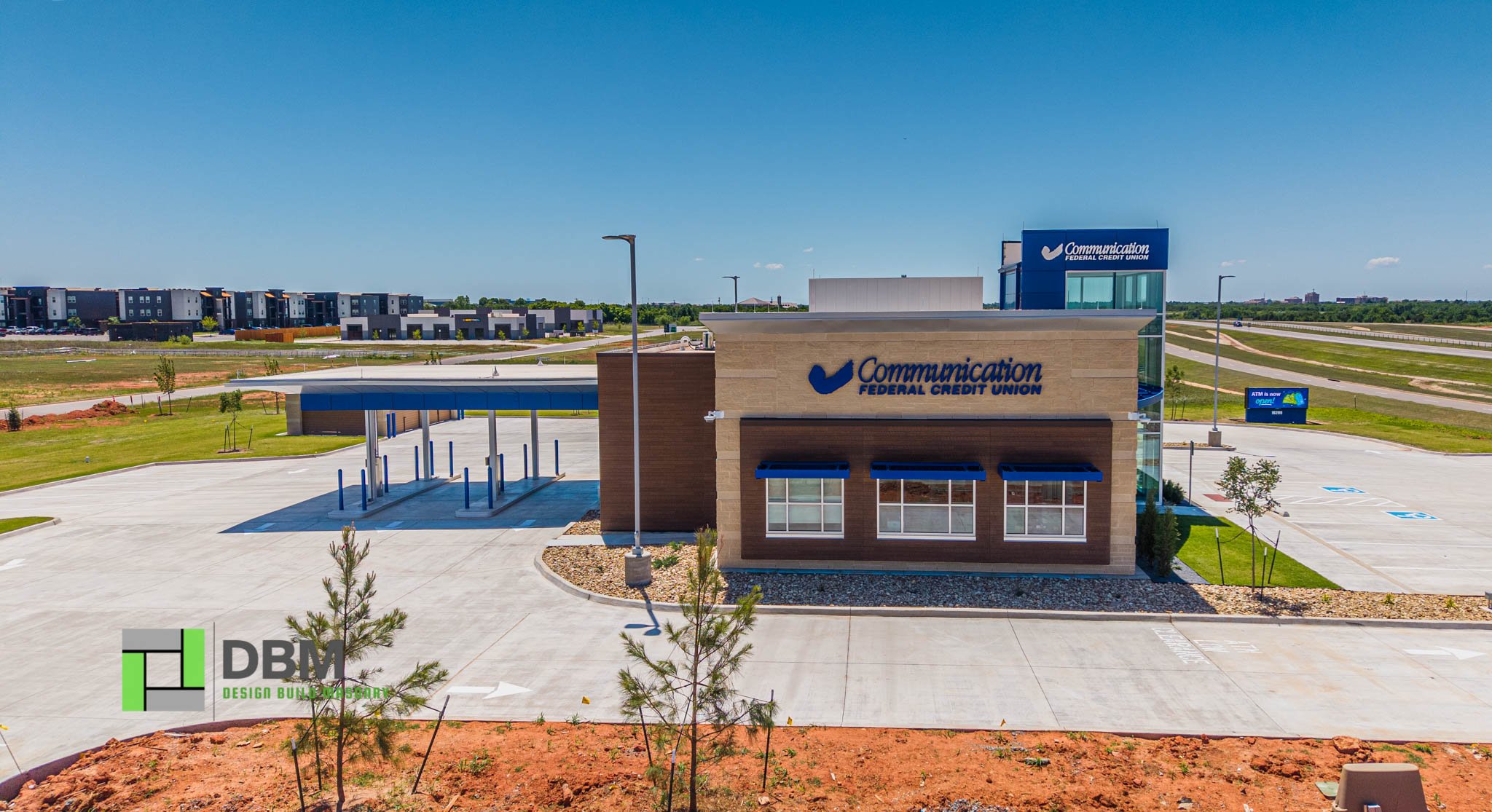 A modern building with the logo and name of Communication Federal Credit Union, situated in a parking lot with a bicycle rack, surrounded by green landscaping, and a backdrop of residential buildings and open sky.