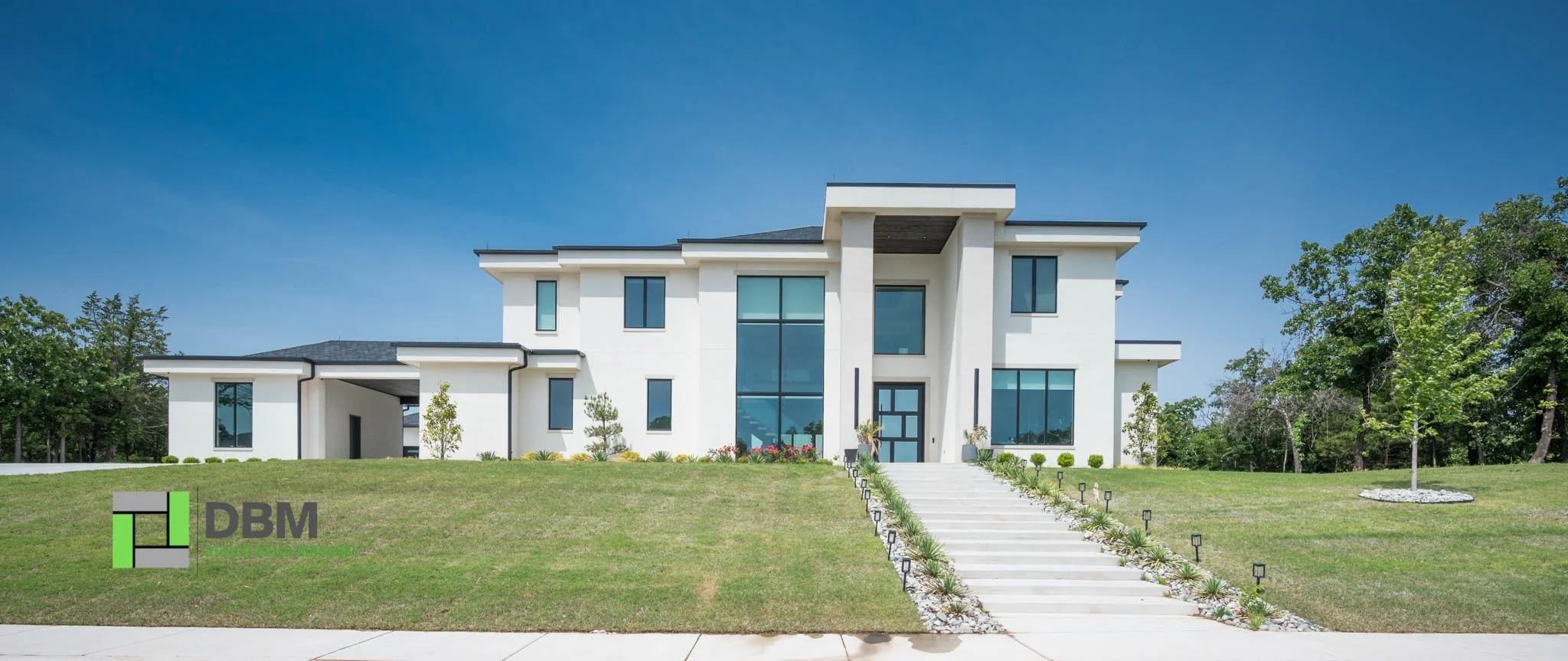 Modern white house with large windows, a staircase leading to the front door, surrounded by green lawn and trees, under a clear blue sky.
