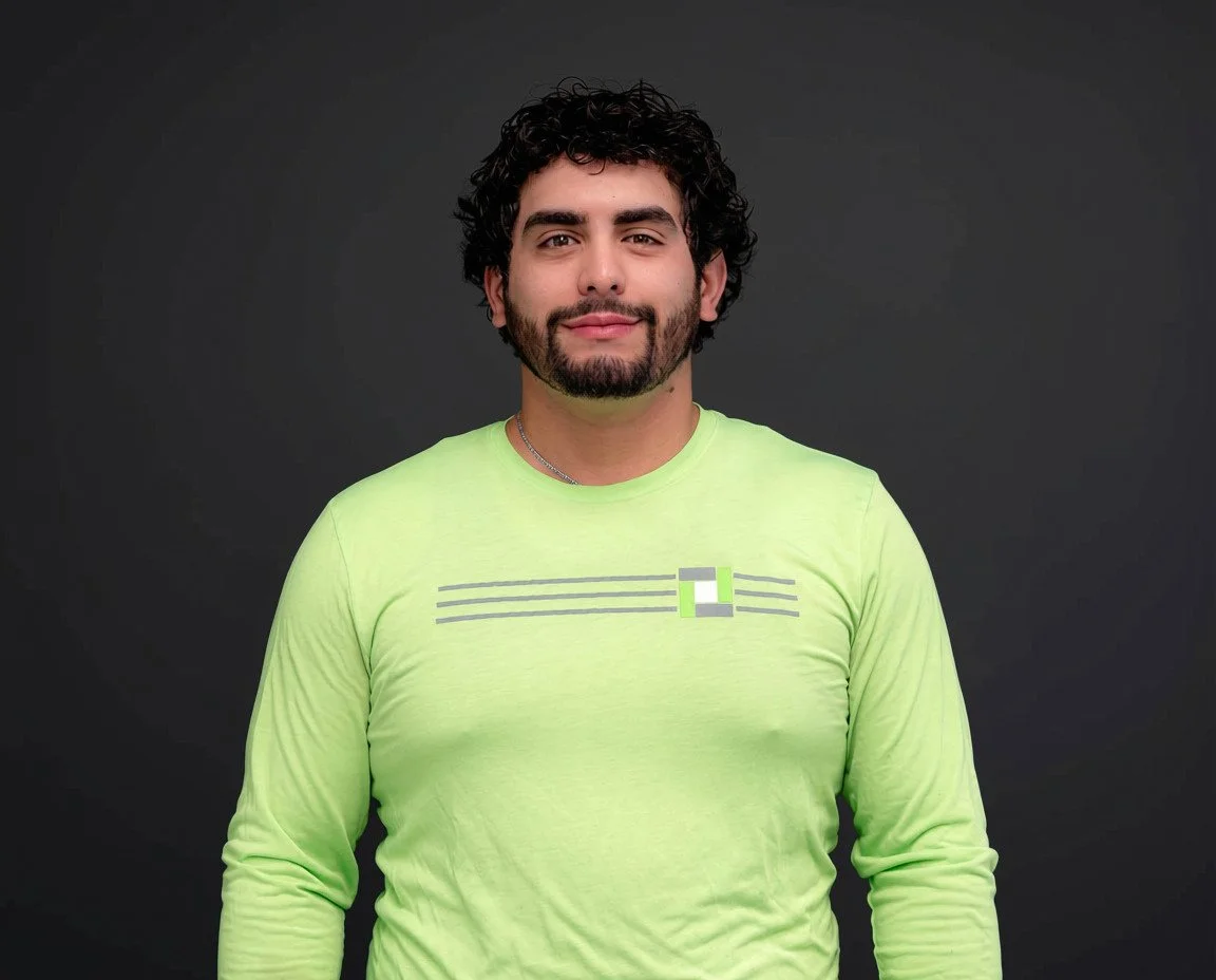 A man with dark, curly hair and a beard, smiling slightly, wearing a neon yellow T-shirt with a geometric design.